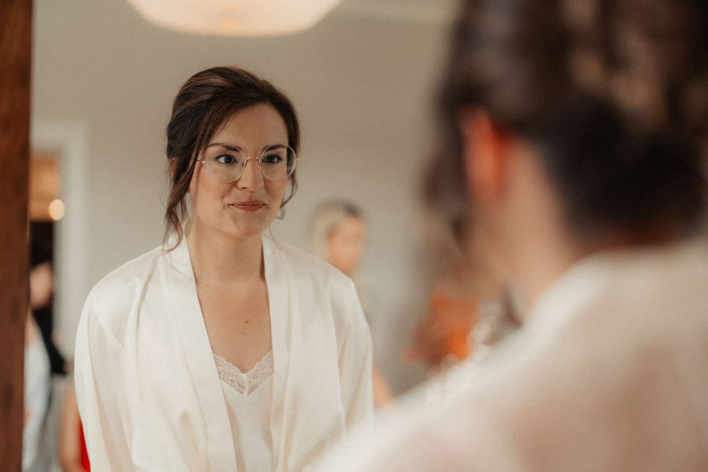 Bride in white robe smiles during morning wedding preparations in soft lighting.