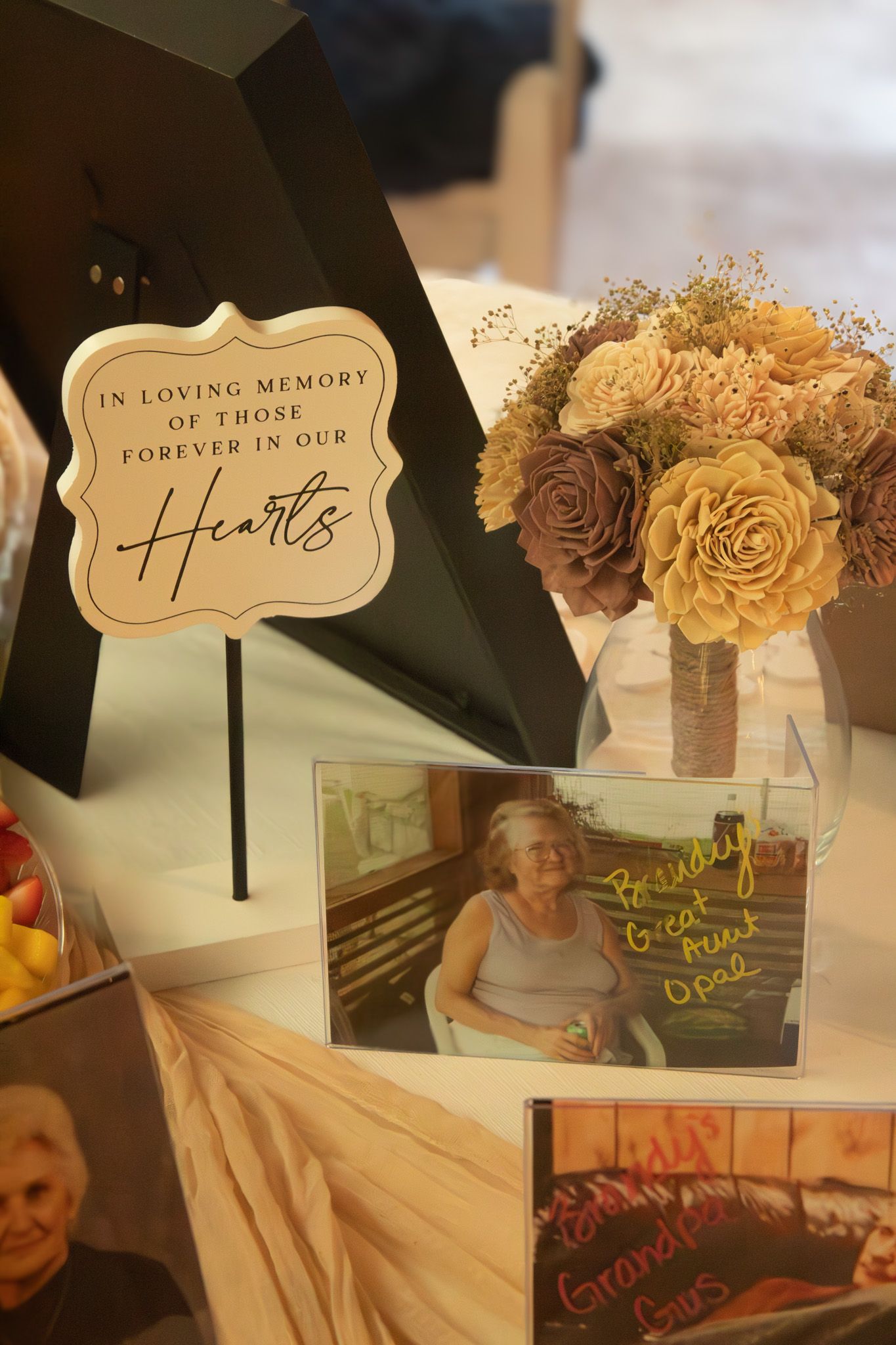 Memorial display with photographs and decorative signs reading 'In Loving Memory' and 'Forever in Our Hearts' with floral accents.