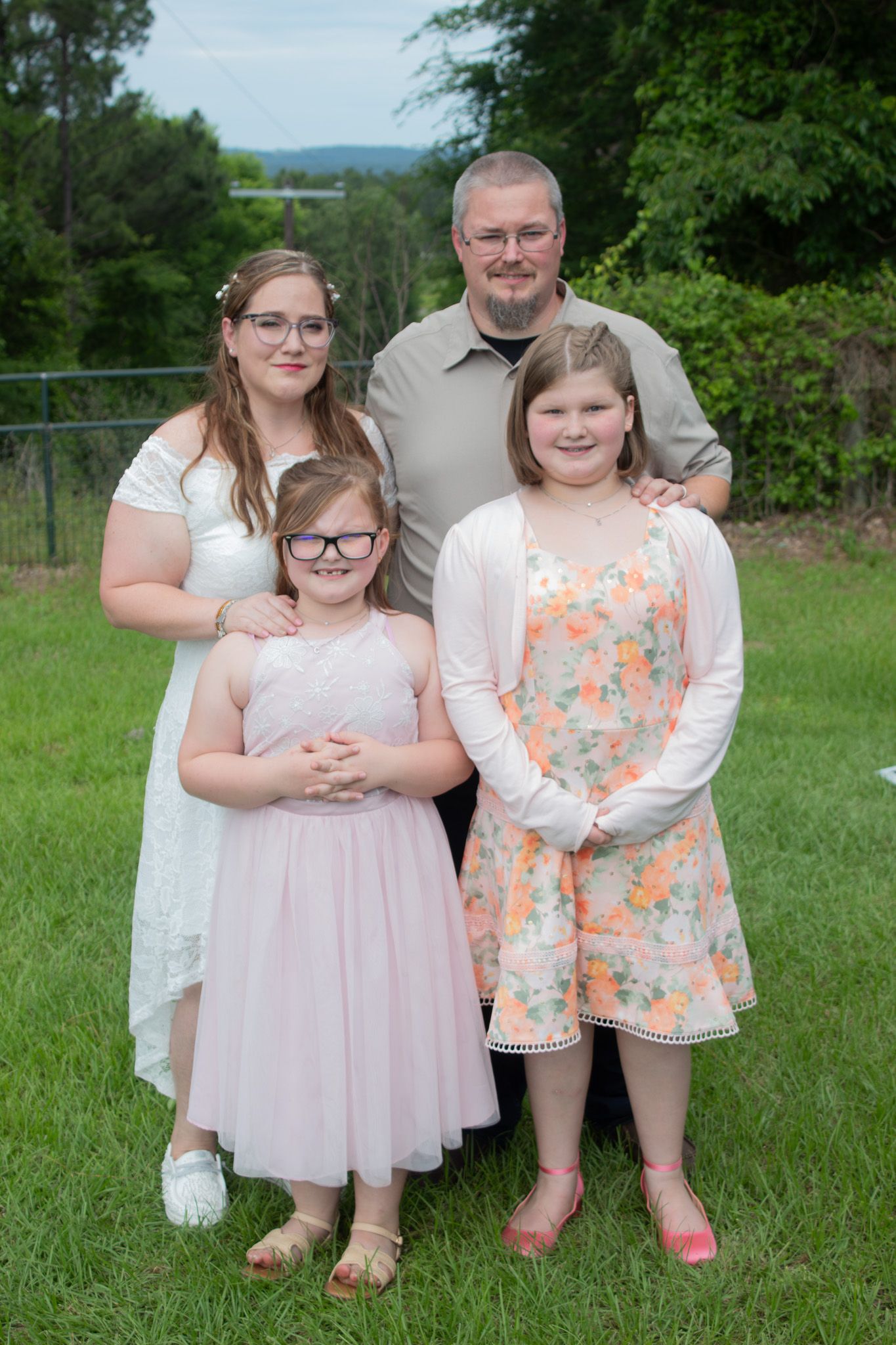 A family of four poses together outdoors on a grassy lawn with trees and greenery in the background.