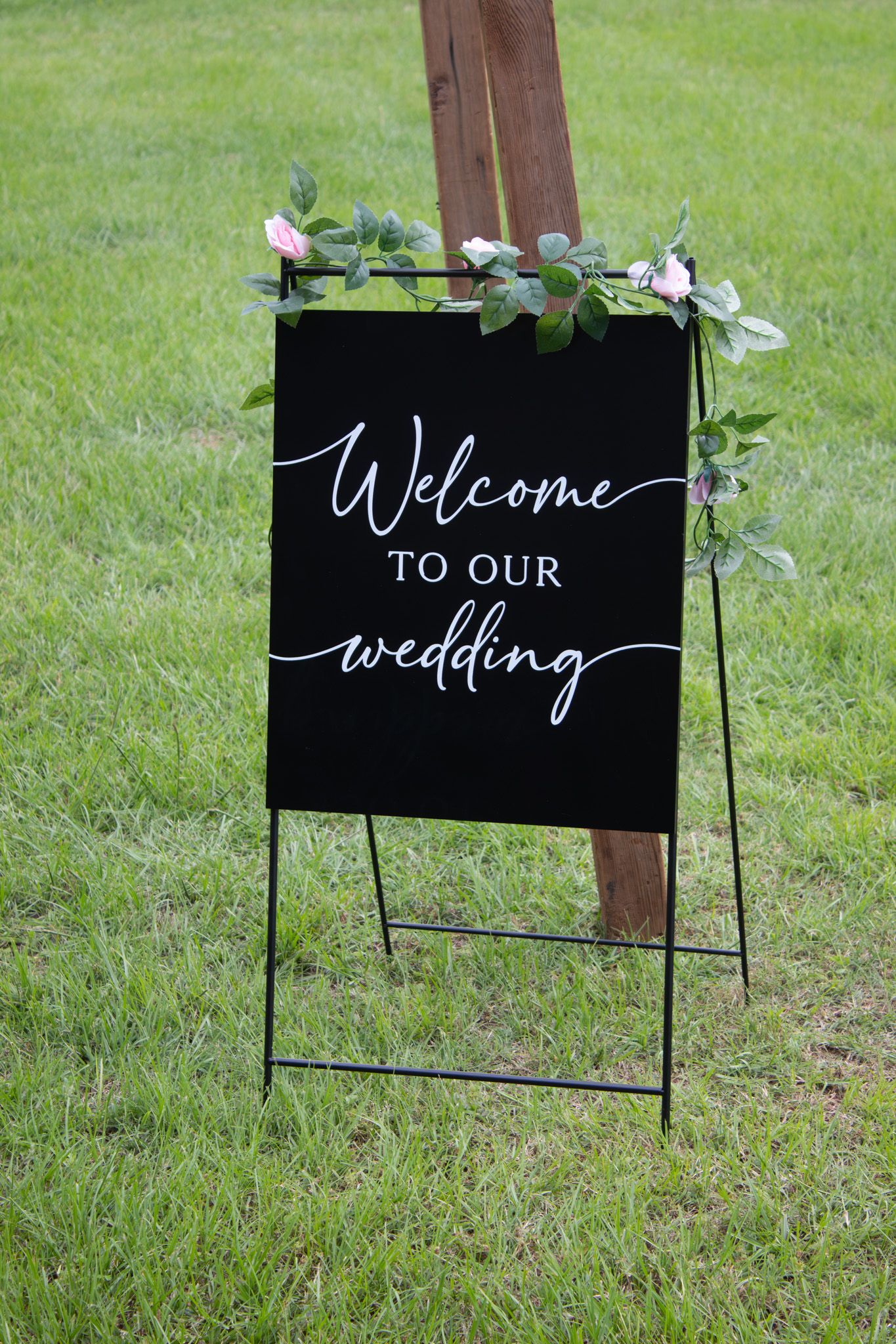 A black welcome sign with white calligraphy text reading 'Welcome to our wedding' stands on grass with decorative greenery.