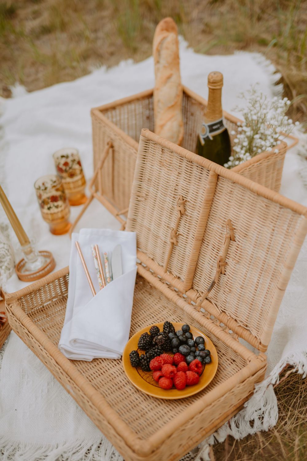 A picnic setup with a wicker basket, bread, champagne, berries on a plate, and glasses on a white cloth.