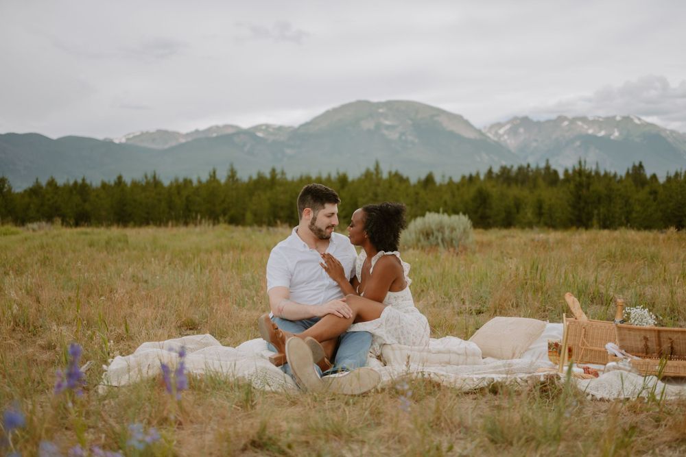 A couple sitting on a blanket having a picnic in a scenic meadow with mountains and a forest in the background.