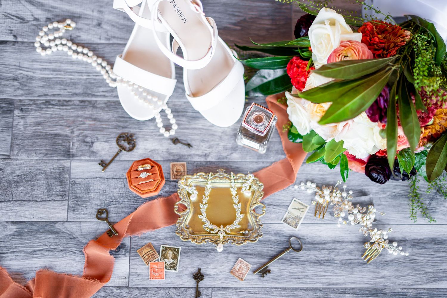 Wedding details flatlay with white shoes, pearls, and floral arrangements on gray wooden surface.