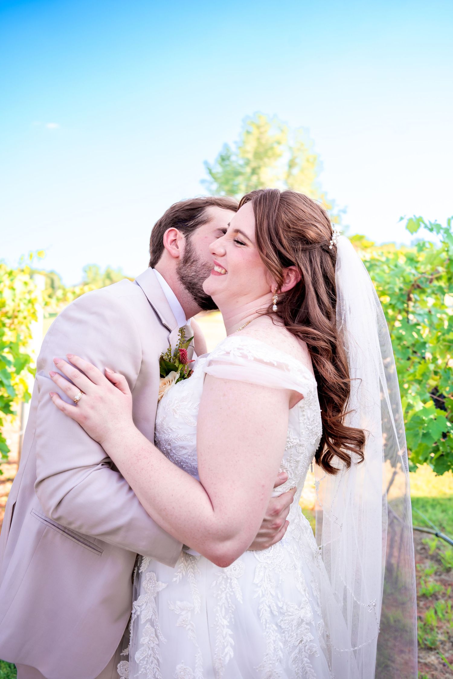 A romantic wedding couple embraces outdoors on their special day with natural sunlight and greenery in the background.