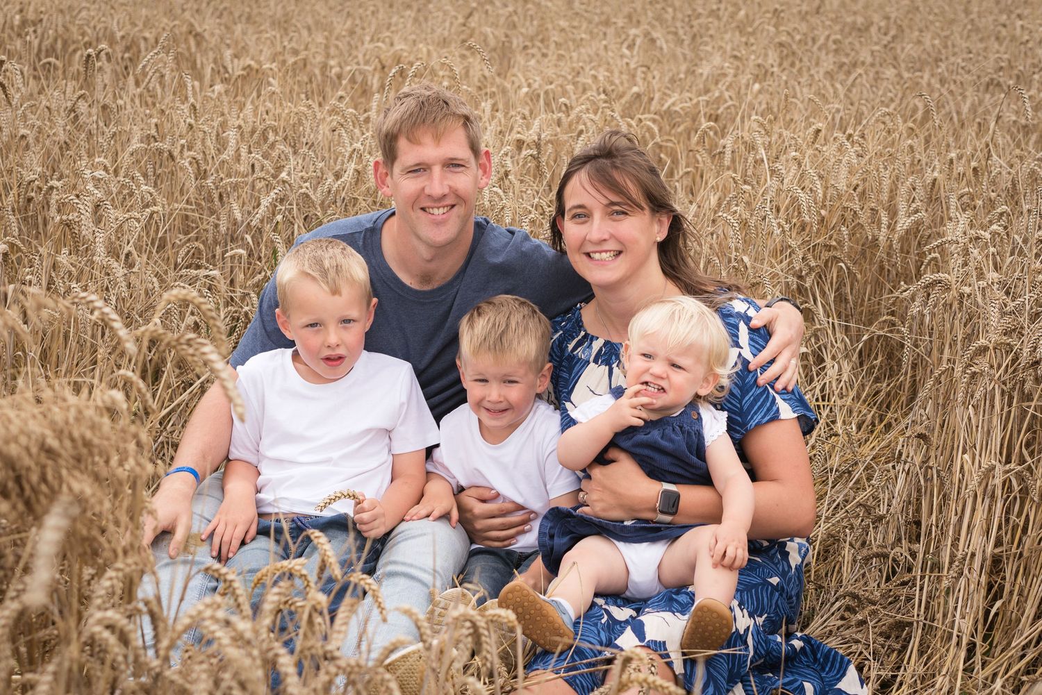 A family portrait with 3 small children, and parents, sat down in a field of wheat, all having a big cuddle.