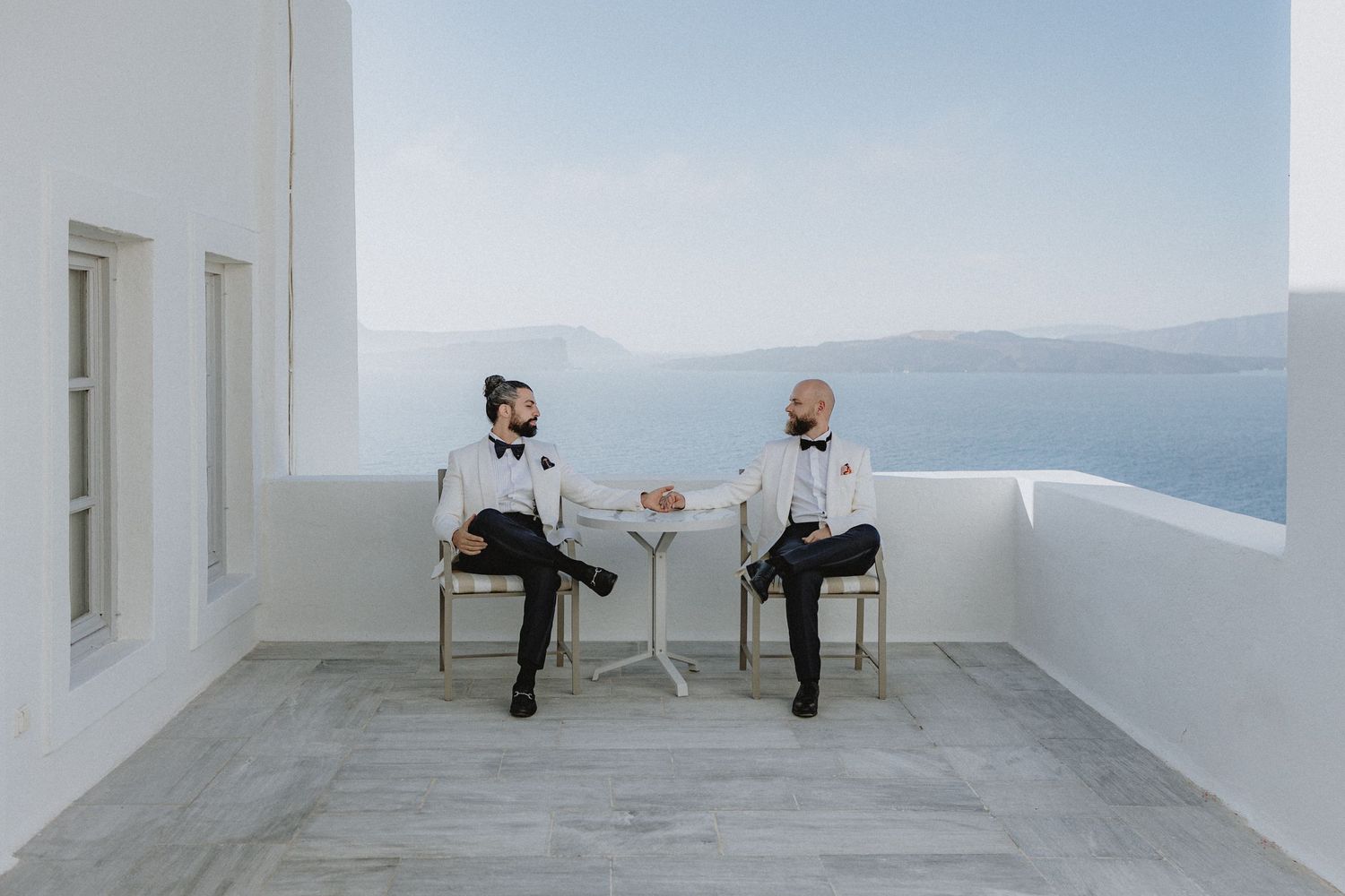 Two grooms in tuxedos with white coats sitting at a small table holding hands in Santorini