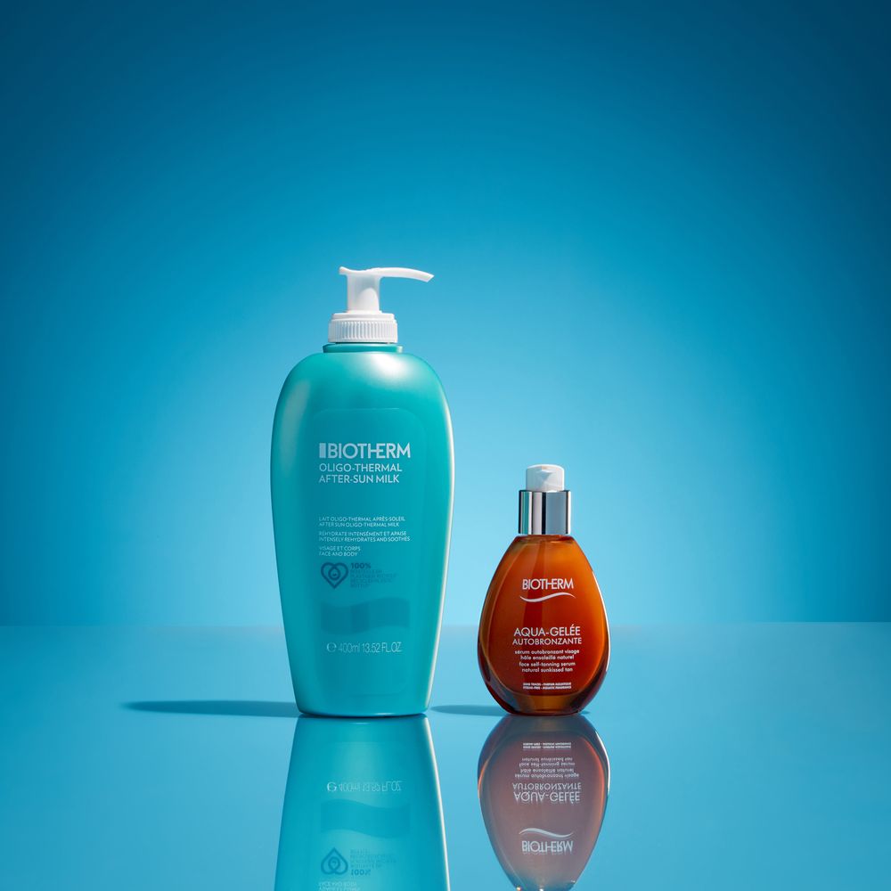 Two skincare bottles, one turquoise pump dispenser and one amber glass bottle, reflected on blue background surface.