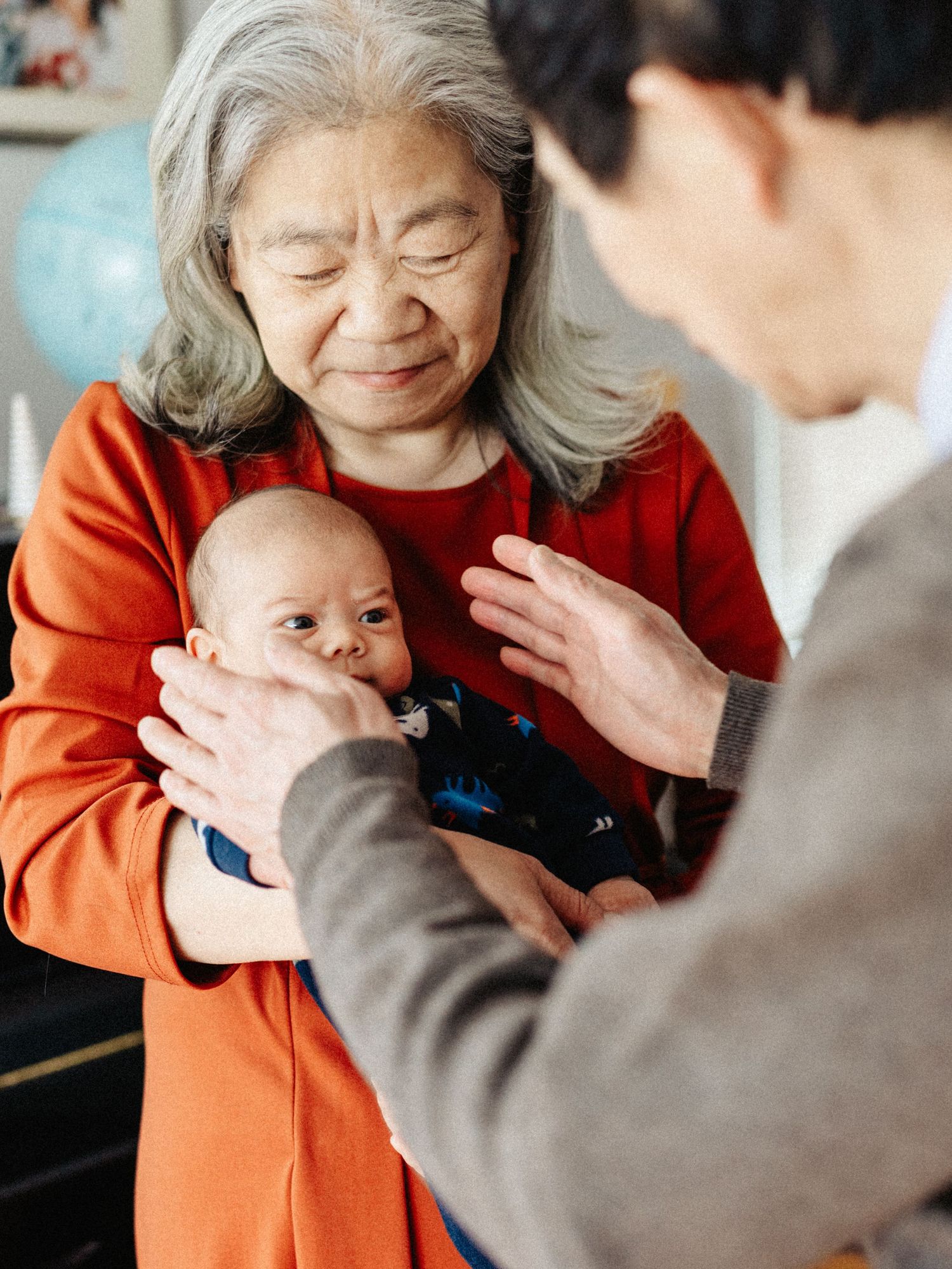 Senior grandmother in orange sweater cradles newborn baby while family gathers around.