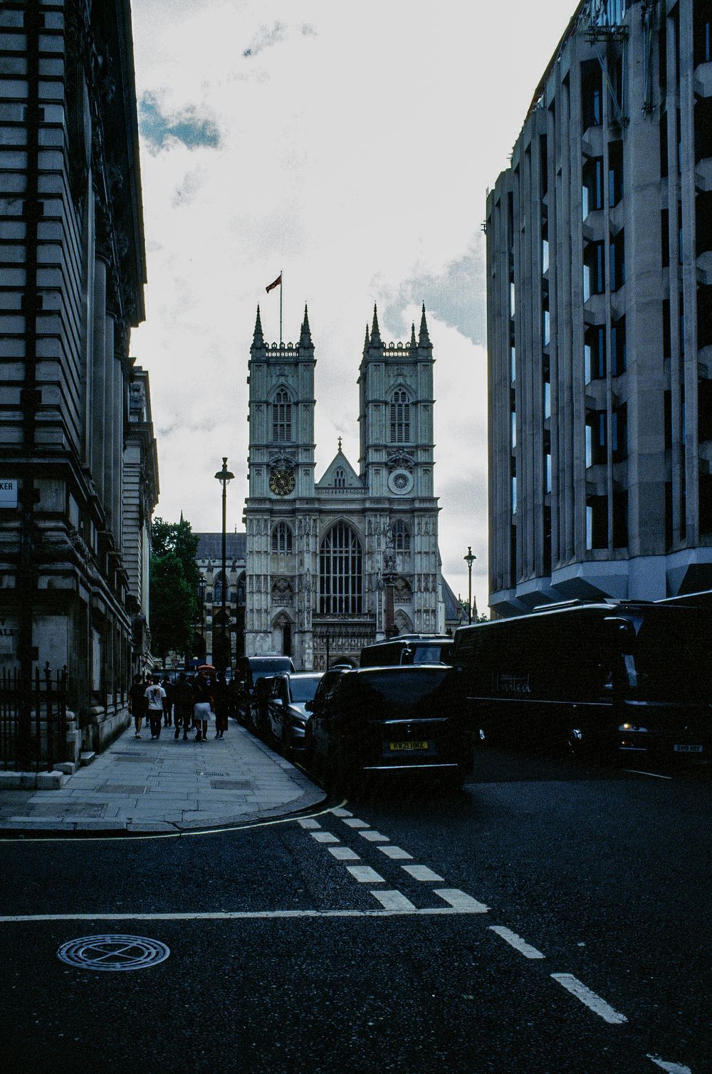 Westminster Abbey's Gothic towers rise between modern buildings on a moody London street.