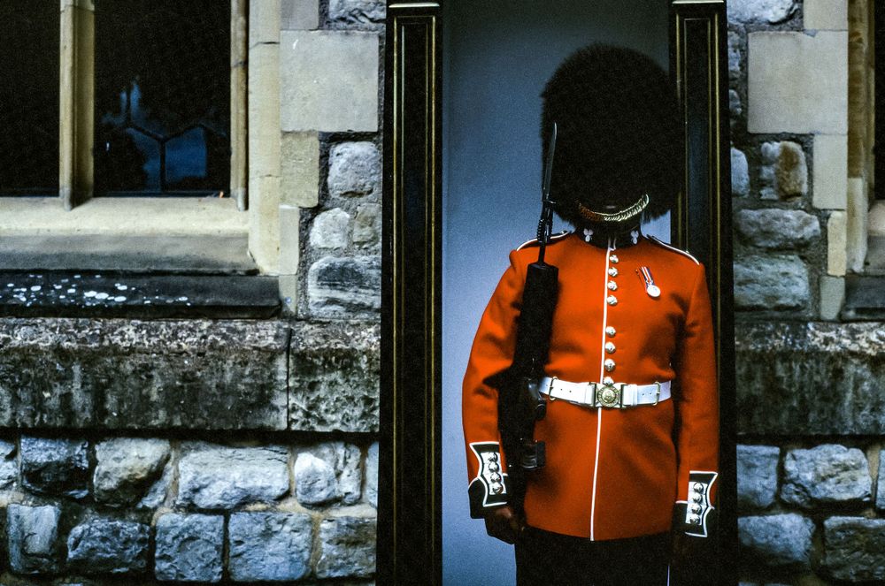 Guard in traditional red uniform stands at attention in stone doorway.