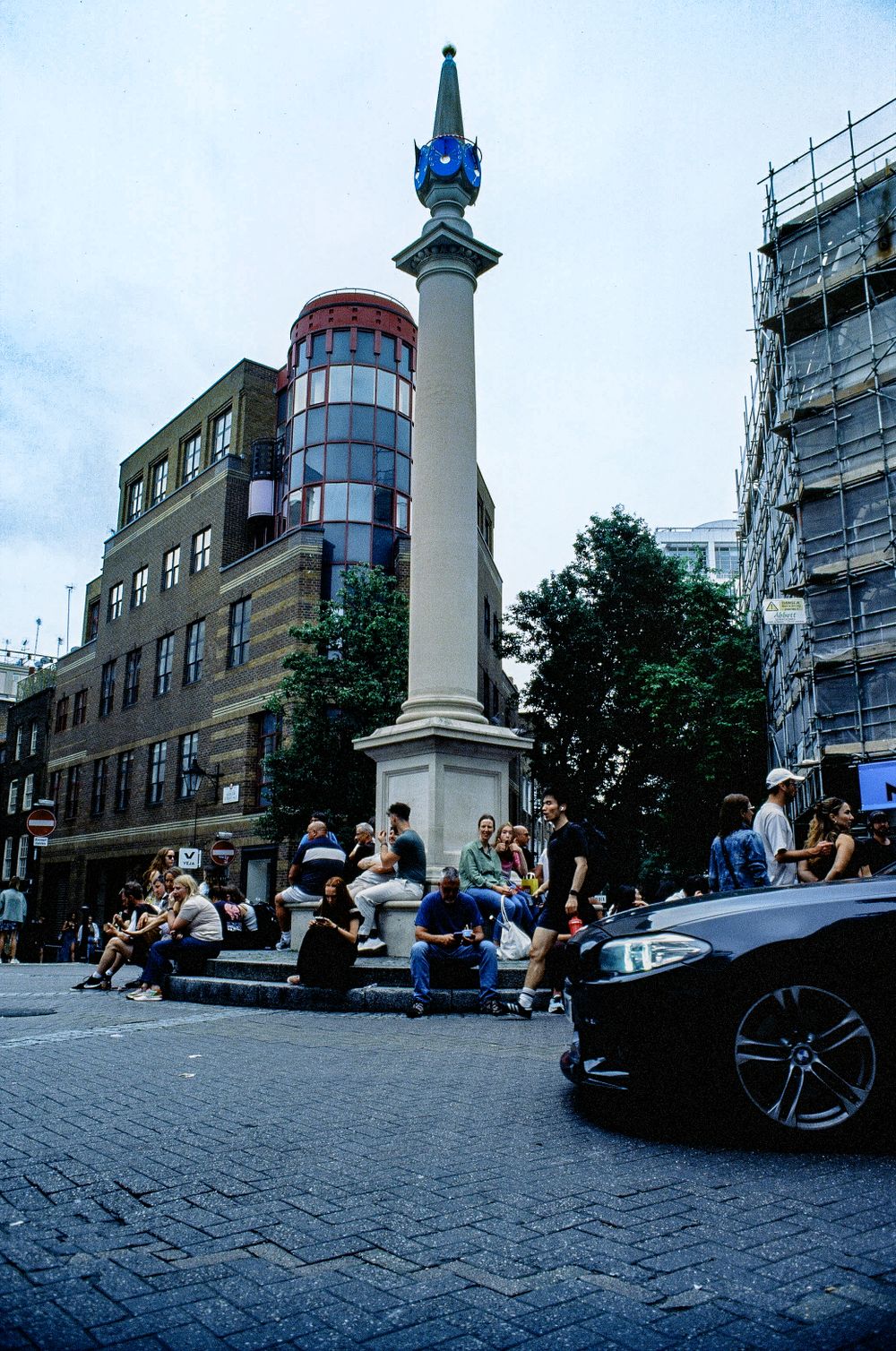 People gathering around a tall ornate column monument on a cobblestone street in an urban setting.