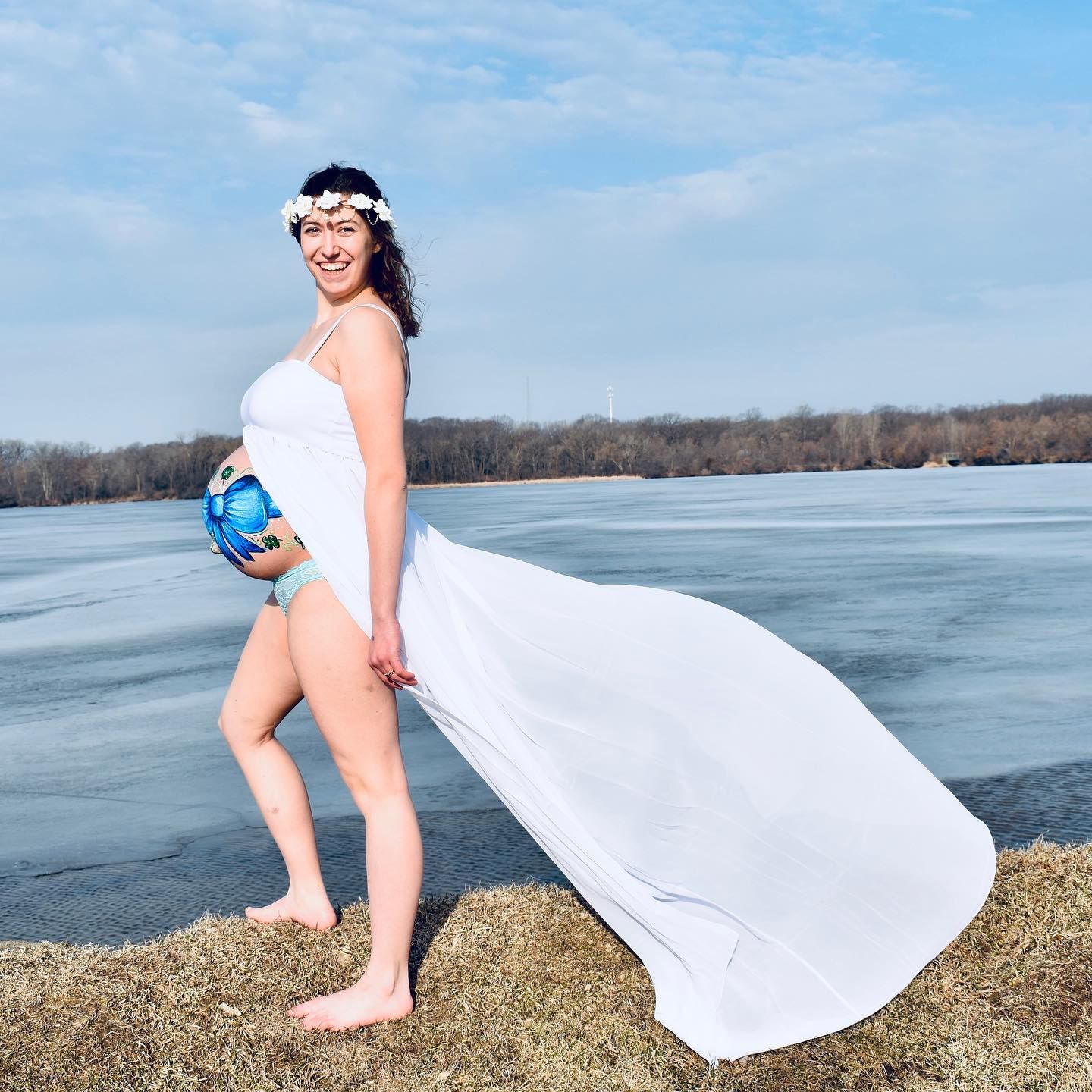 Woman in flowy white dress poses playfully on frozen lake during maternity photoshoot.