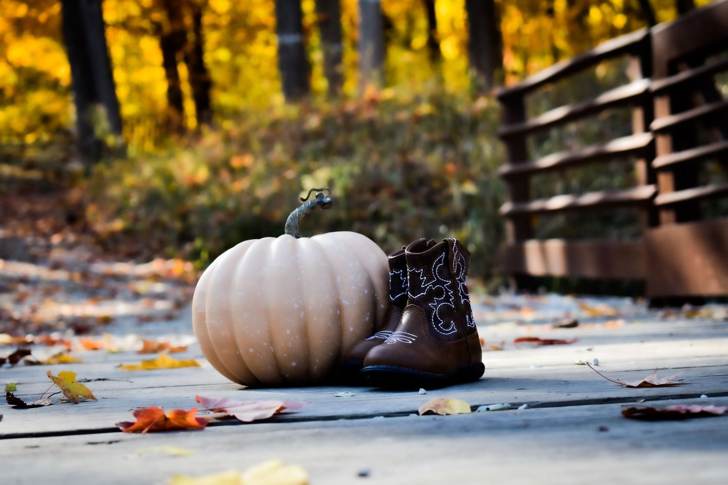 A white pumpkin and brown leather boots arranged on a wooden deck with fall leaves.