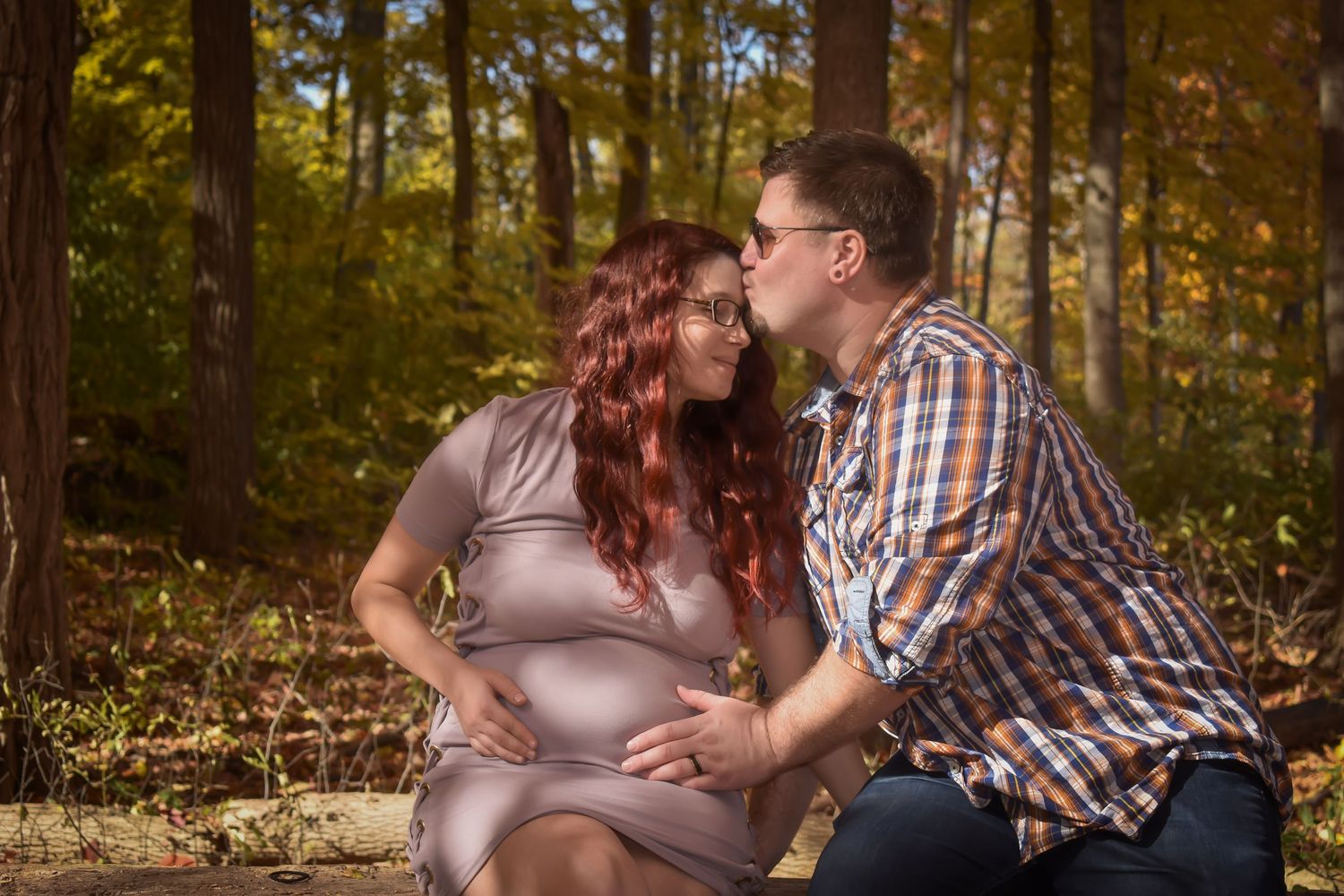 A romantic autumn maternity photo session in a wooded setting with golden fall foliage in the background.