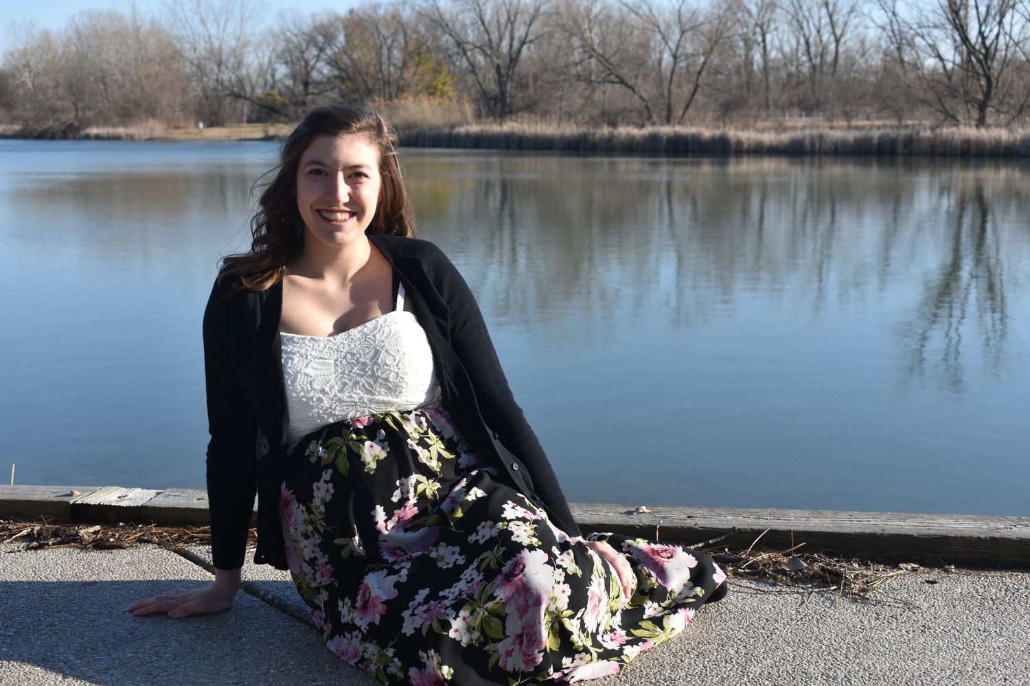 Woman in floral dress sits by lakeside wearing black cardigan.