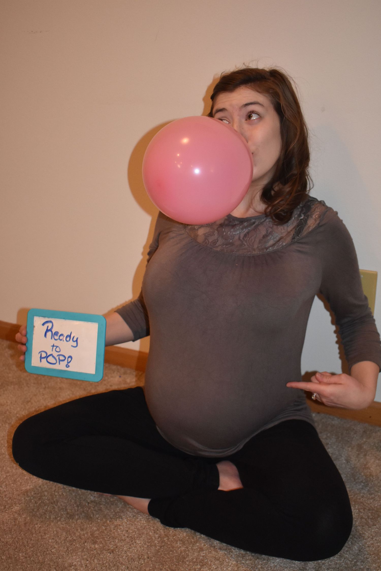 Pregnant woman in brown shirt blowing up pink balloon while sitting cross-legged on carpet.