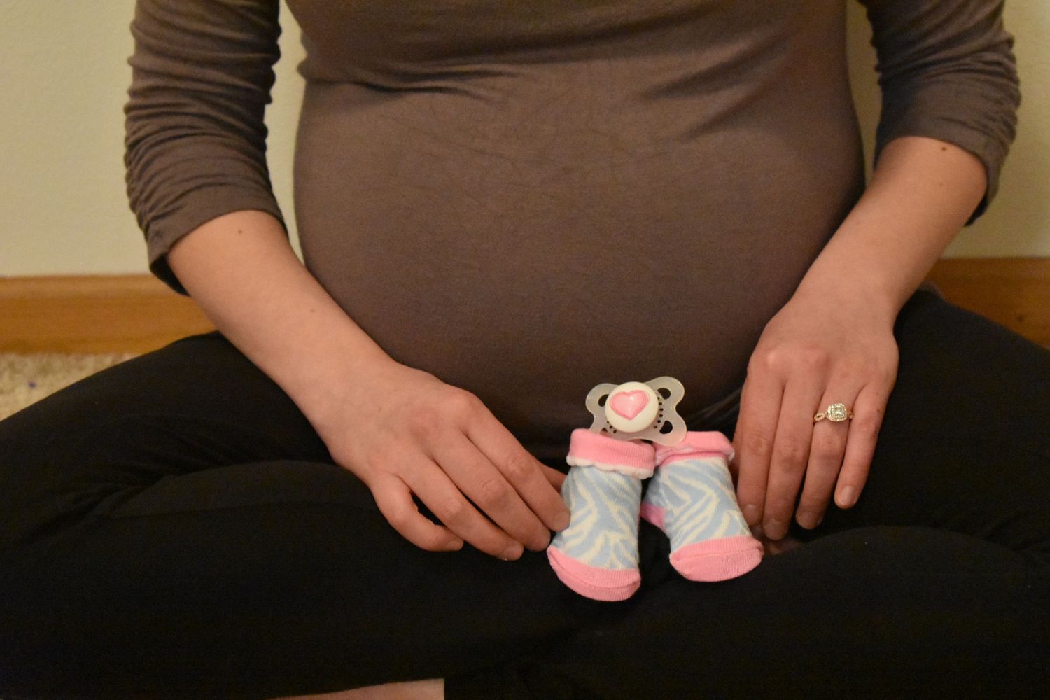 Small pink and gray baby booties held over pregnant belly in brown sweater.