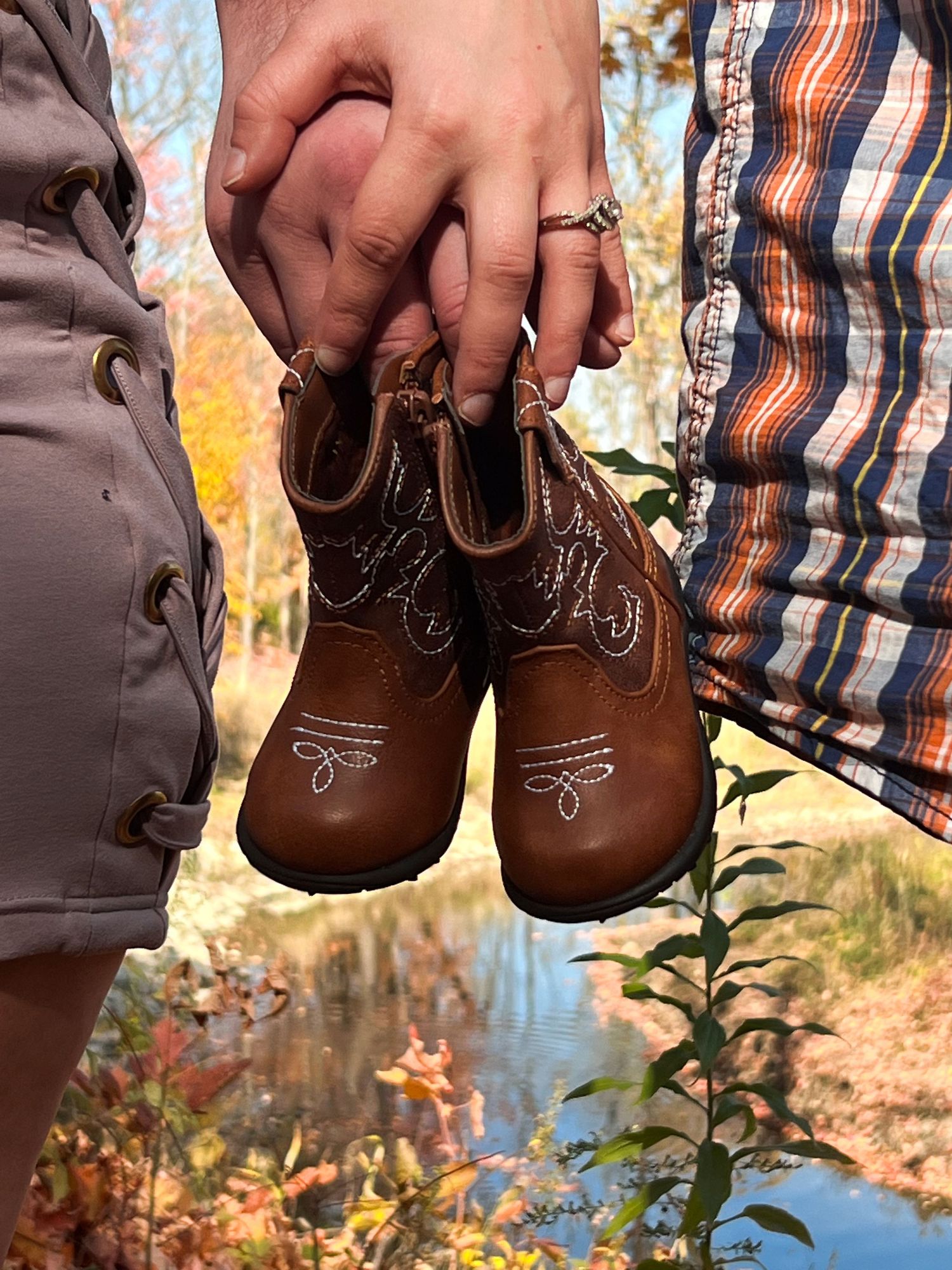 Hands holding tiny brown baby boots against an autumn forest backdrop.