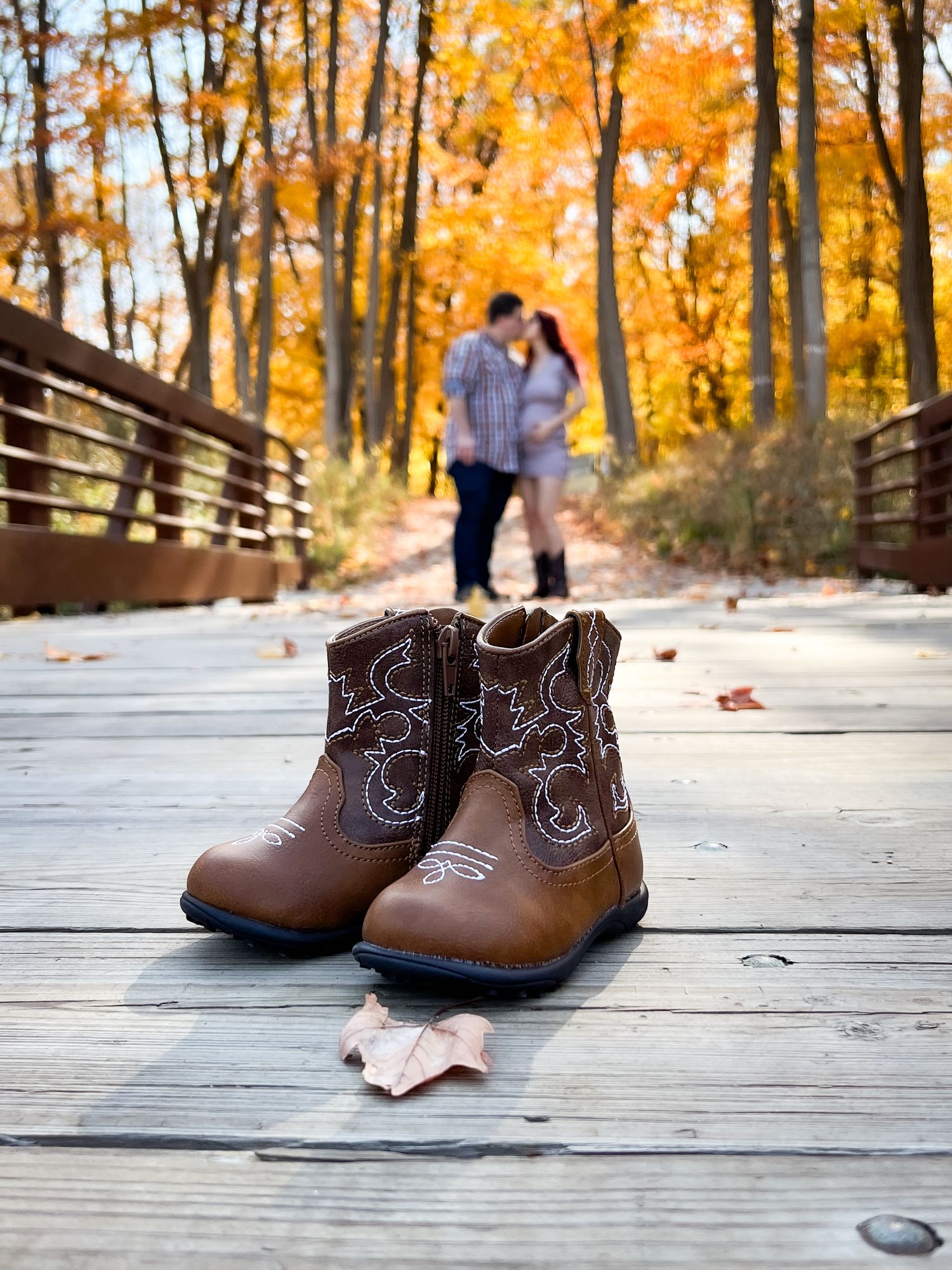 Small brown cowboy boots on wooden bridge with fall foliage in background.