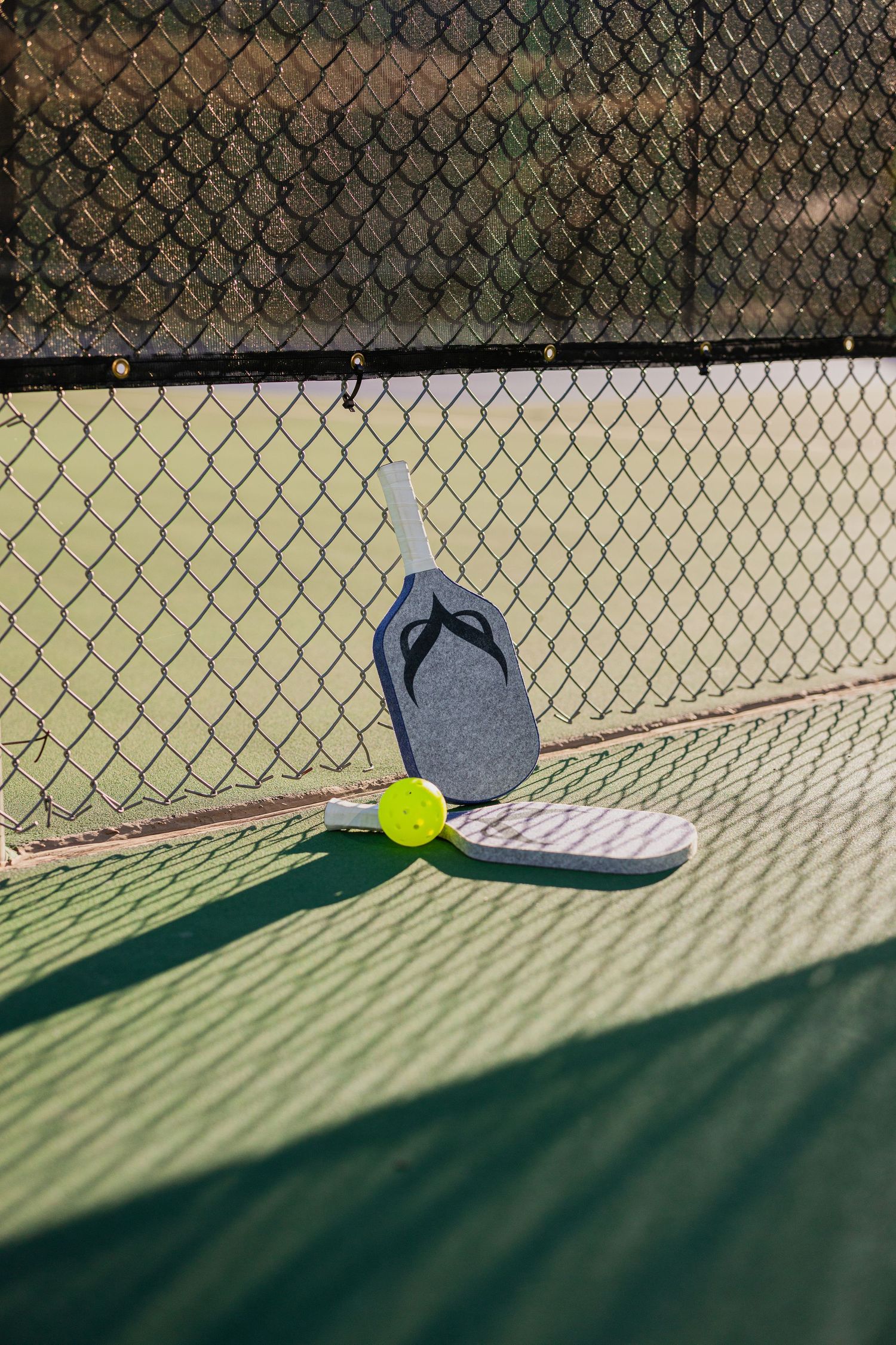 Pickleball paddle and yellow ball casting shadows on a tennis court with chain-link fence.