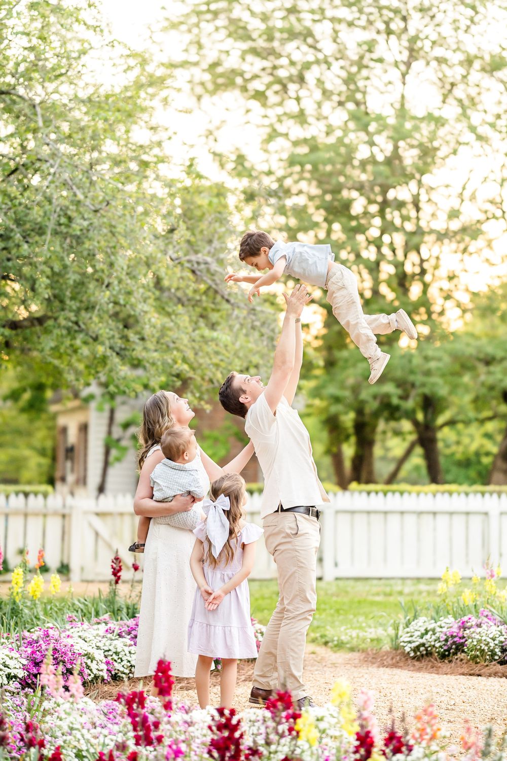 Photograph of a family of five interacting lovingly and playfully while dad tosses one child in the air in a light and airy garden in CW.