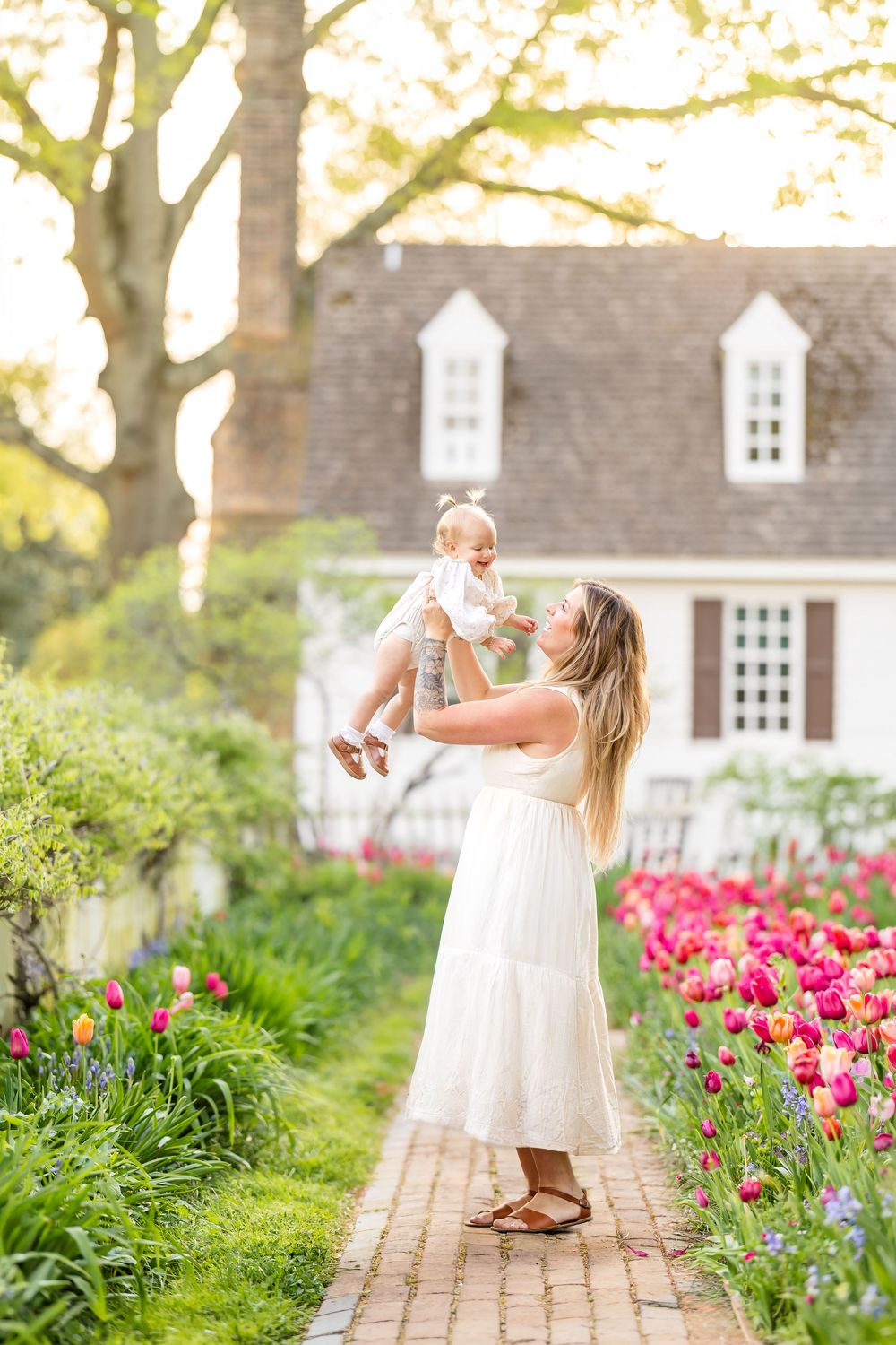 Golden light photography of a mother in a white dress lifting a toddler in the air affectionately while in the Williamsburg tulip garden.