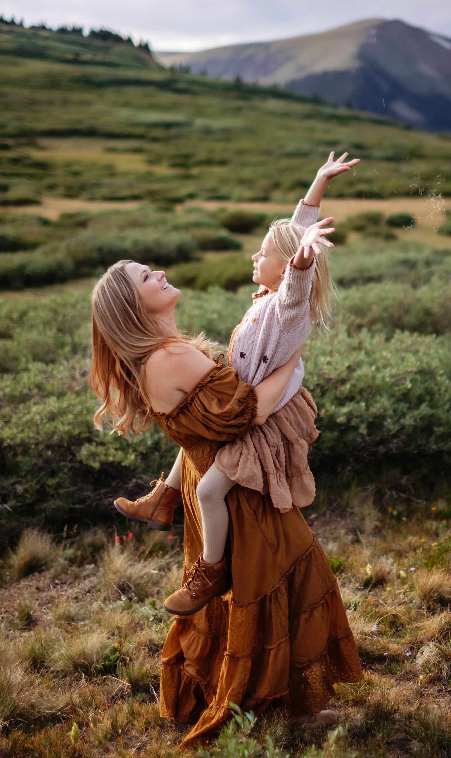 A woman in a flowy brown dress dances freely in the mountain meadow with her arms raised against rolling hills in rocky mountains colorado