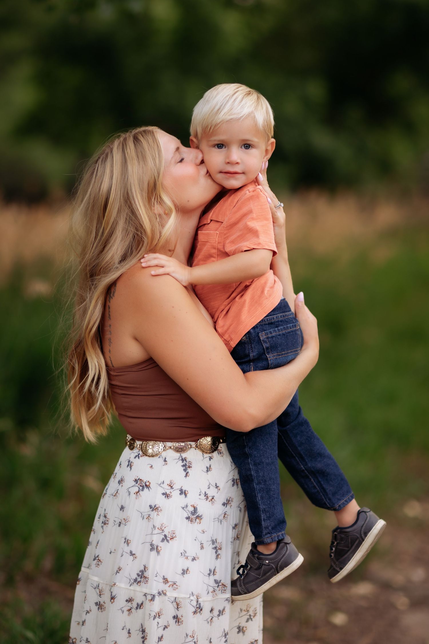 A mother wearing a brown top and floral skirt lovingly embraces and kisses a small child in an orange shirt outdoors in Navarre Florida