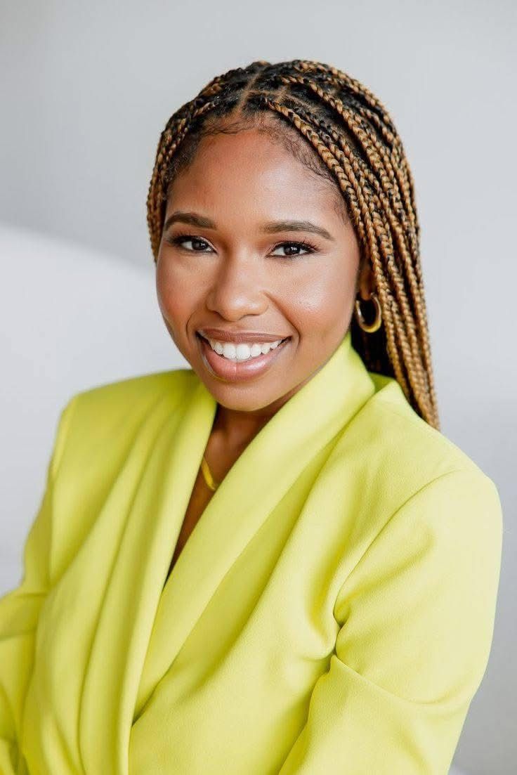 Professional headshot of person in bright yellow blazer with braided hair and radiant smile.