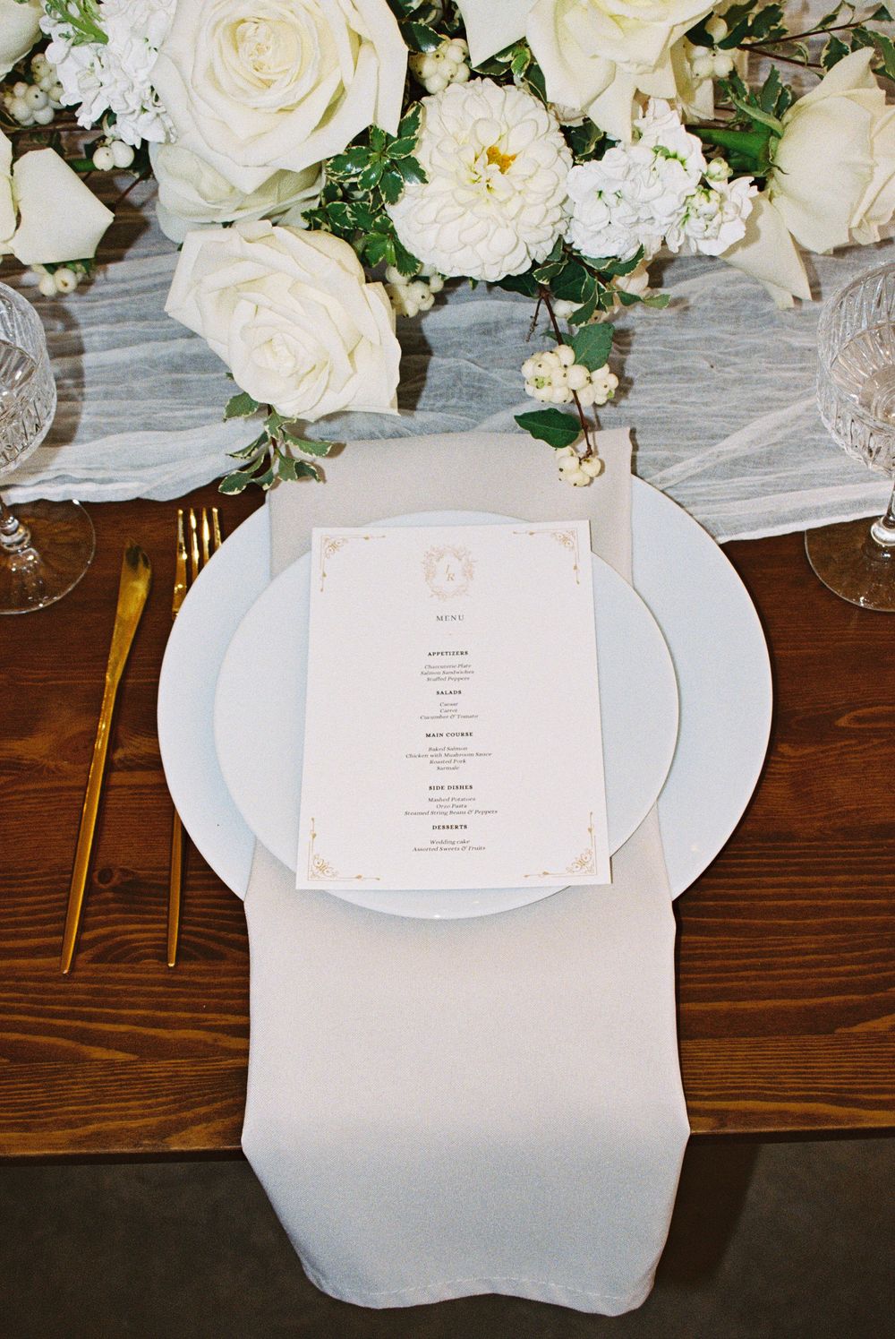 Elegant wedding place setting with white roses, menu card, gold flatware on wood table.