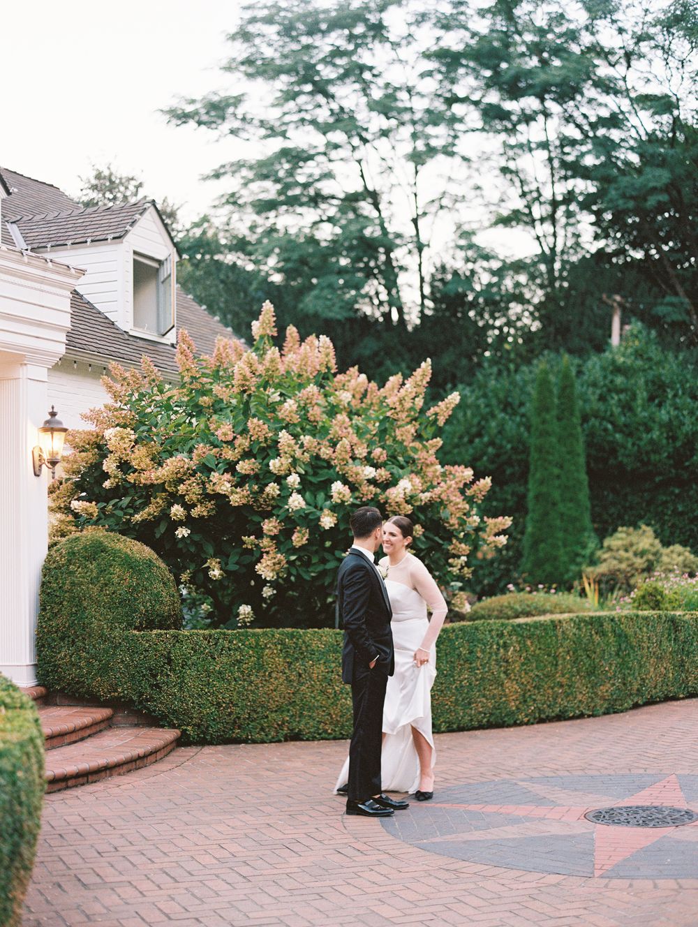 Bride and groom sharing romantic moment in garden at dusk near blooming shrubs.