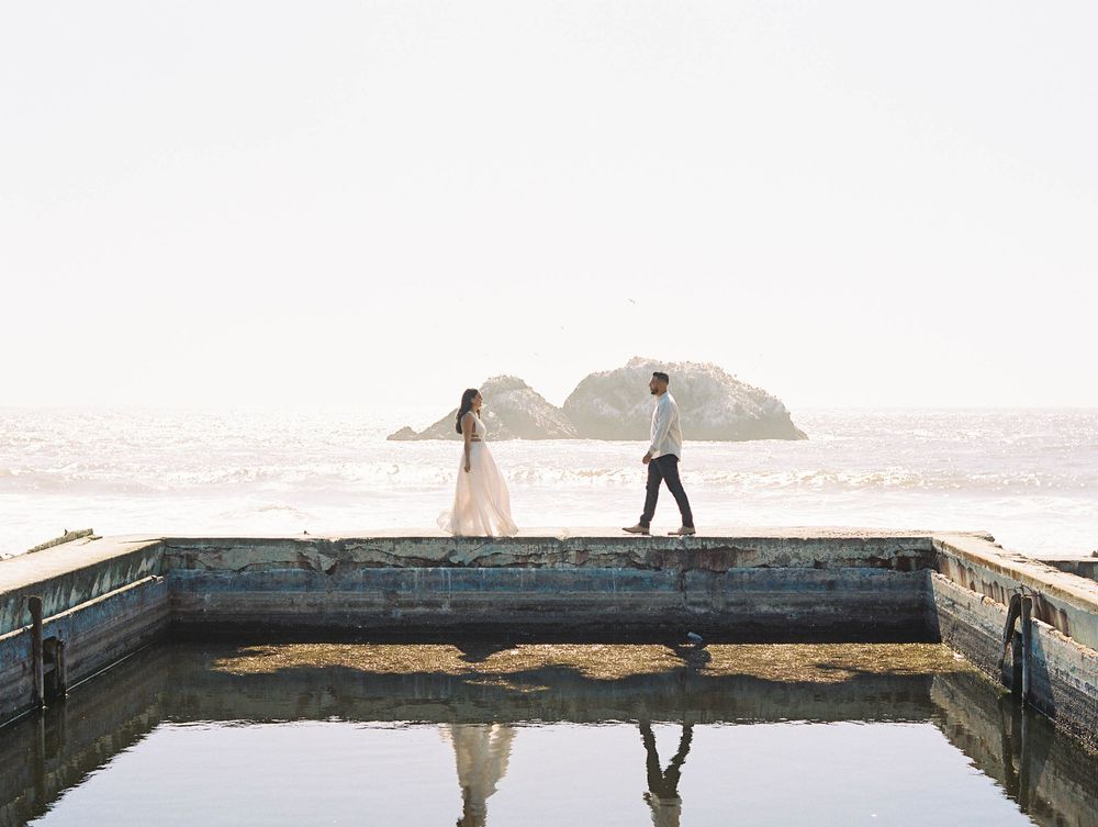 Couple walking along a reflective pool ledge with ocean and rocky island in background.