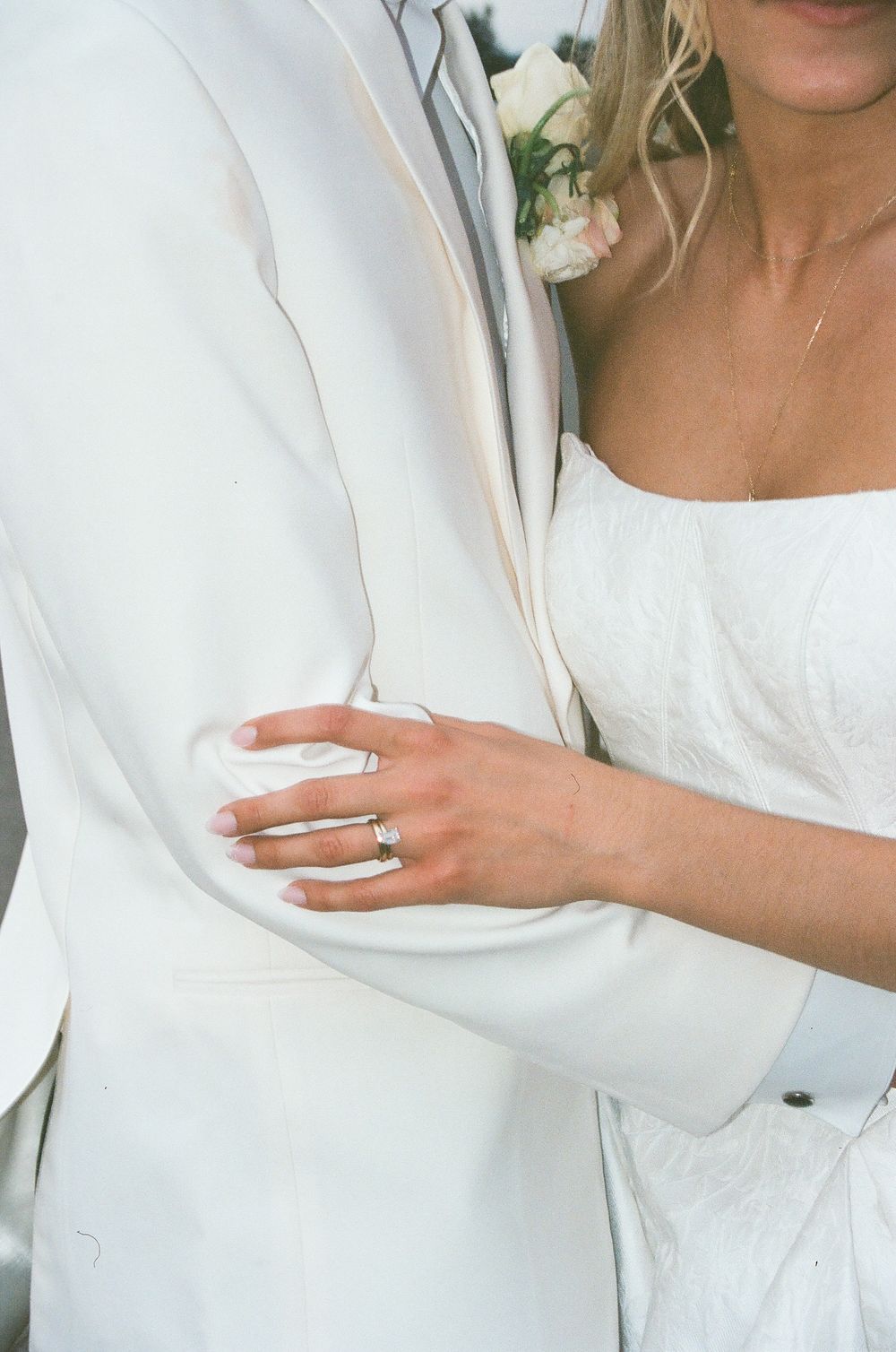 Close-up of bride holding grooms white tuxedo arm showing wedding ring set.