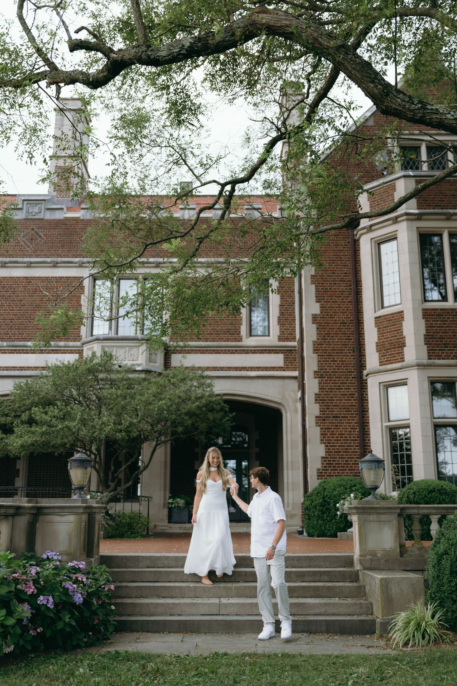 A romantic couple stands on stone steps in front of a grand brick mansion with blooming hydrangeas.