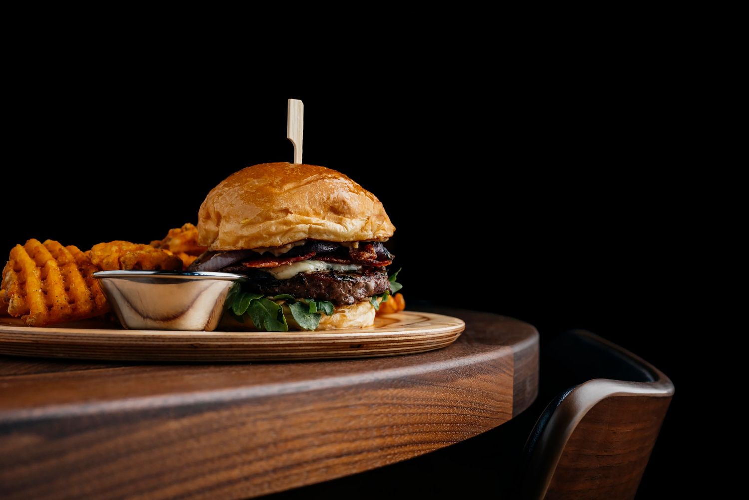 Juicy gourmet burger served on wooden board with waffle fries and dipping sauce against a dark background.