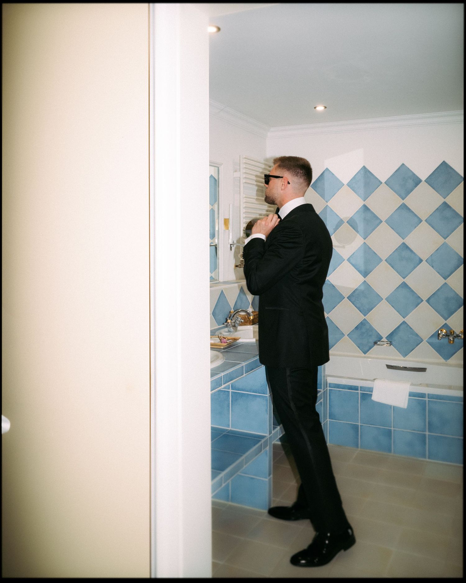 Someone in a black suit adjusts their tie in a bathroom with blue and white checkered tiles.