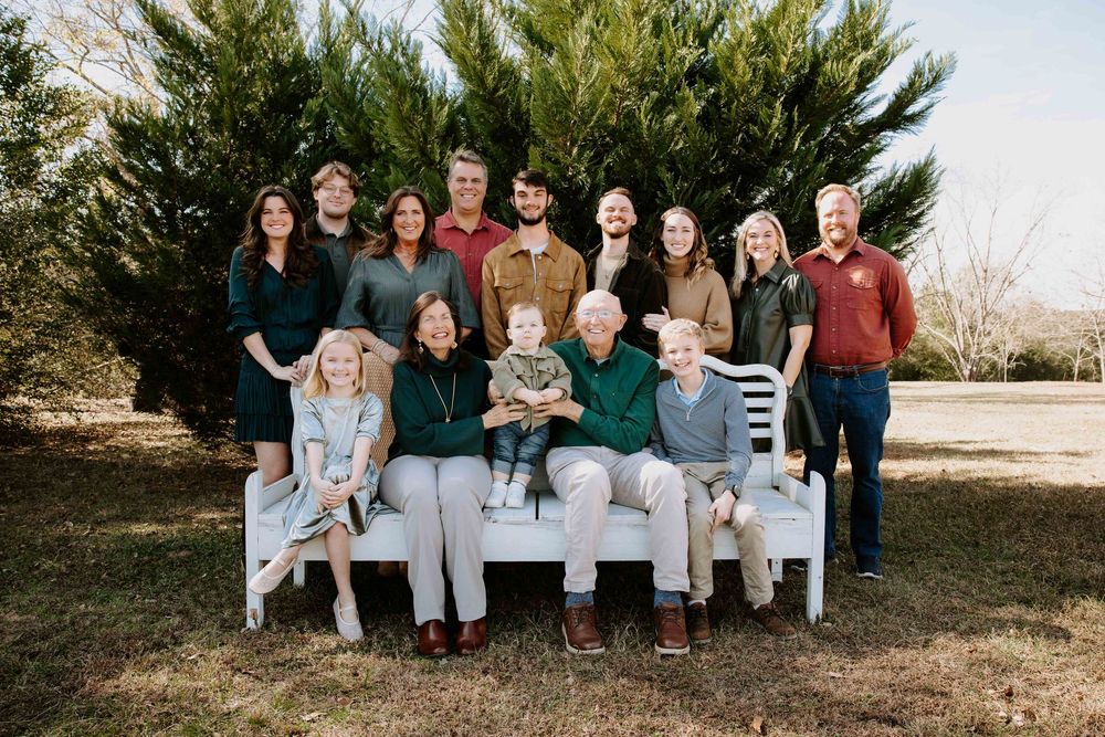 Extended family poses together on white benches outdoors during a sunny autumn photoshoot.