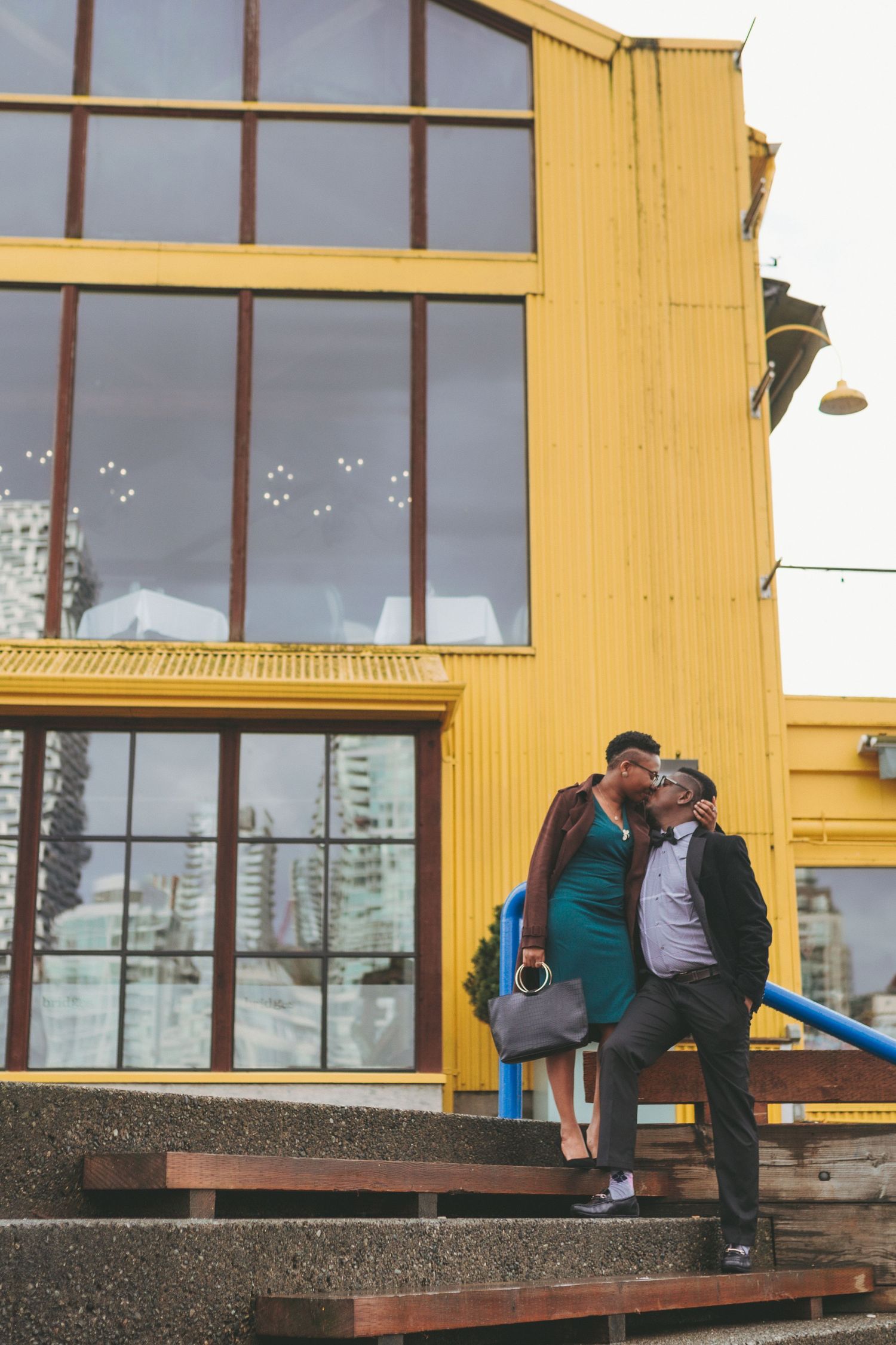 Two figures sit close together on concrete steps against a yellow building with large windows in an urban setting.