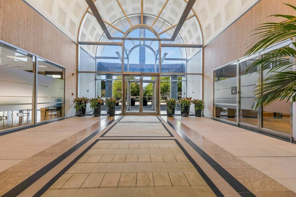 Grand building atrium entrance with arched glass ceiling, marble floors, and lush green potted plants.