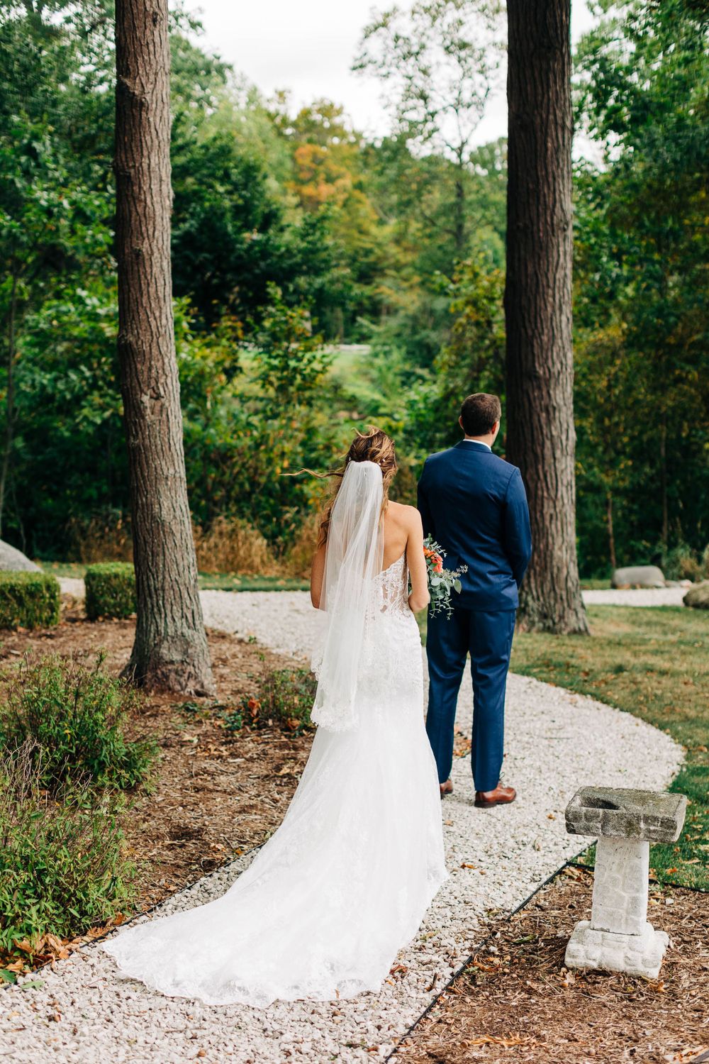 Couple walks down gravel path between tall trees, bride's long white veil trailing behind on their wedding day.