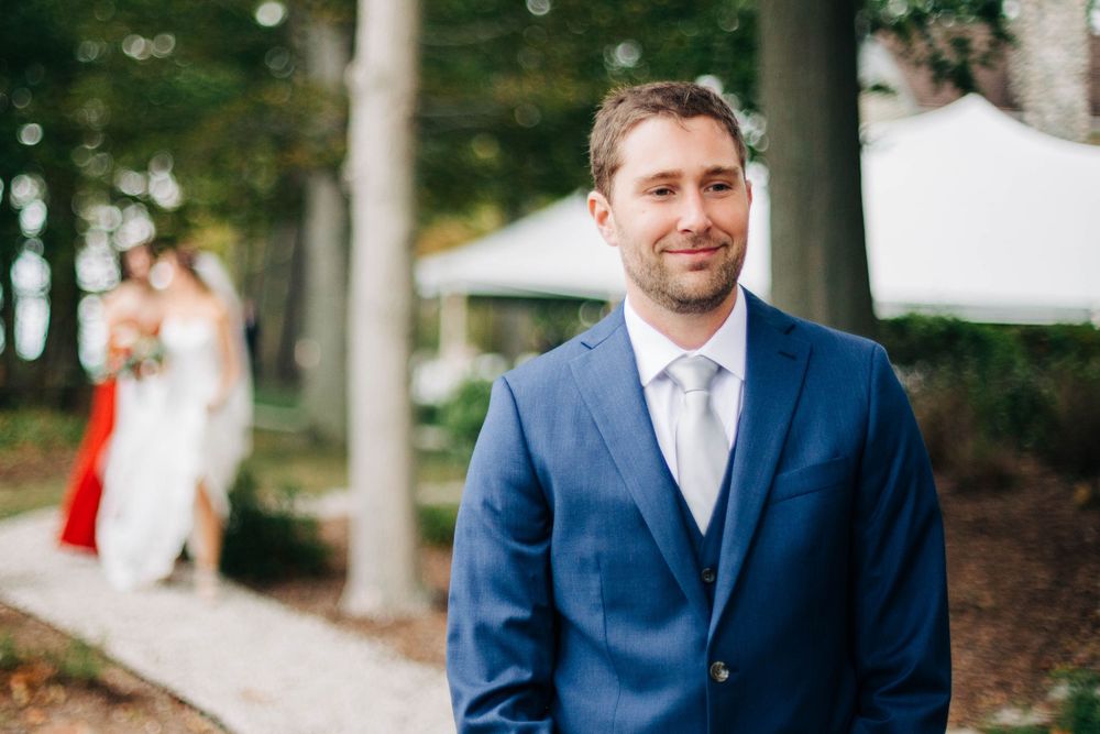 Groom in navy suit stands on wooded path during wedding ceremony.