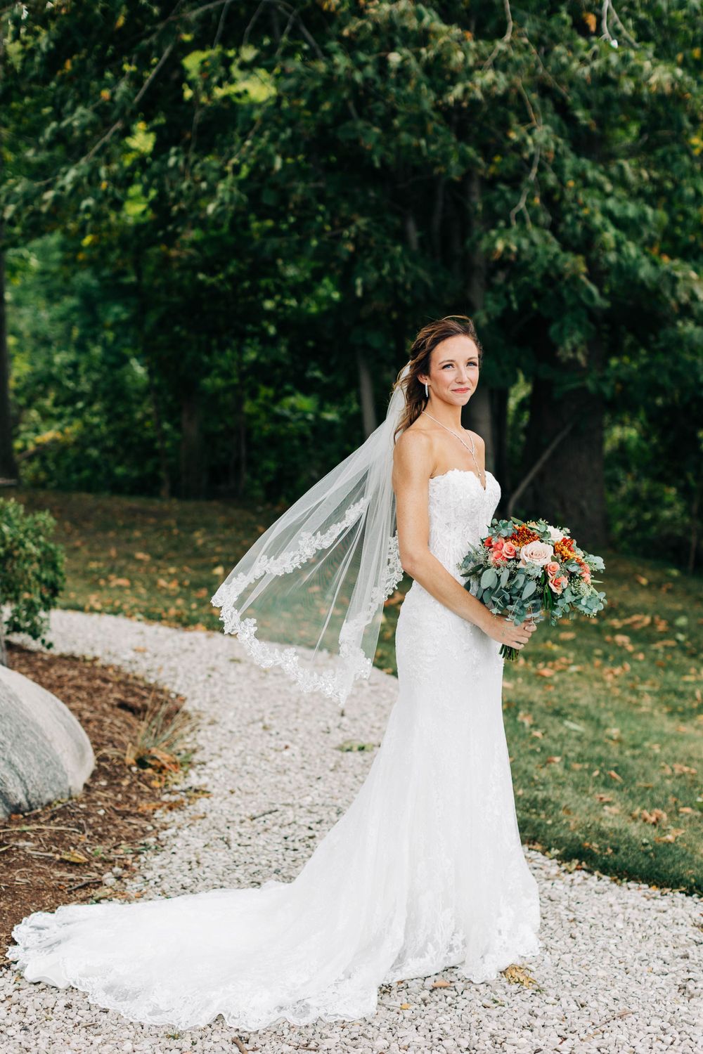 Bride in fitted lace gown poses on garden path with cascading veil and coral bouquet.