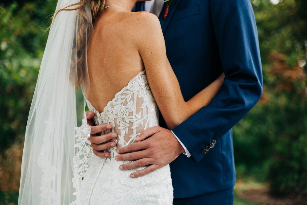 Close-up detail of wedding couple embracing, showing intricate lace dress and navy suit.