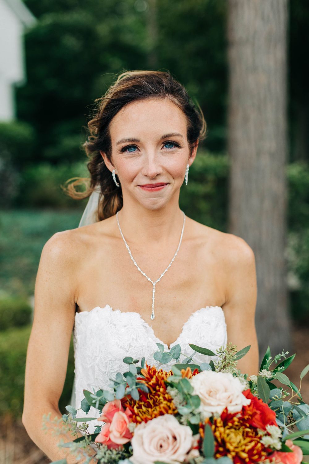 Bride in strapless lace gown holds vibrant autumn bouquet with red and peach flowers in outdoor setting.