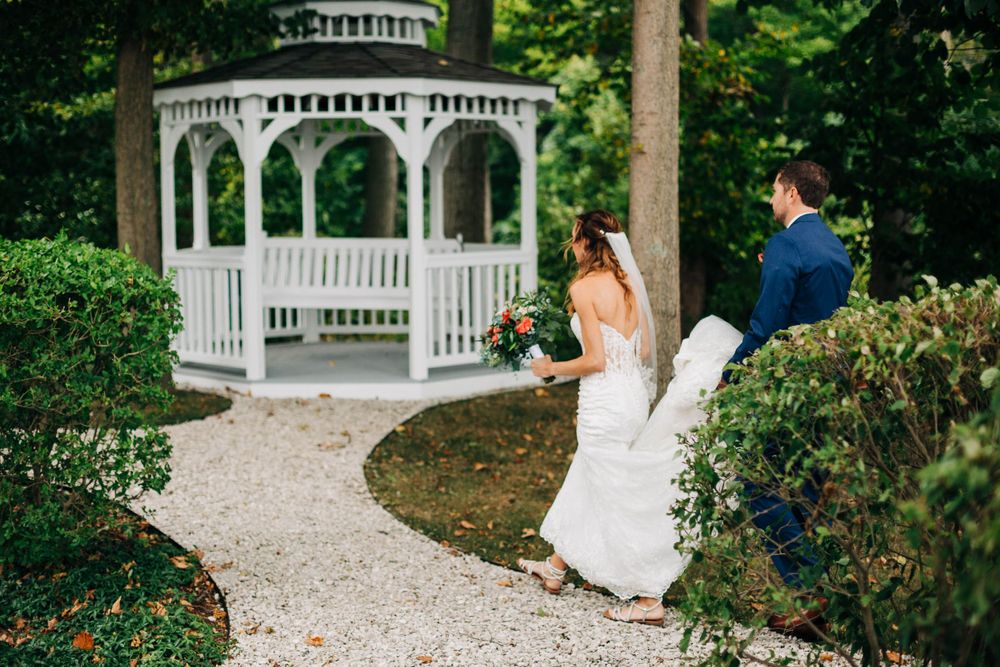Newlyweds walk along garden path towards white gazebo surrounded by lush greenery.