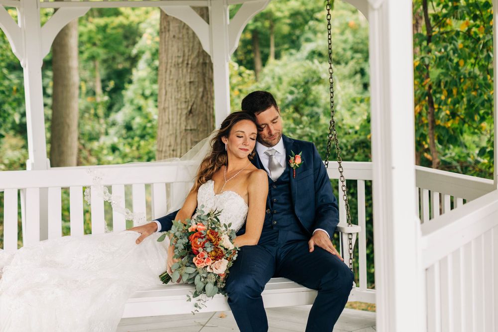 Wedding couple sits on white gazebo porch swing with autumn bouquet and navy suit.