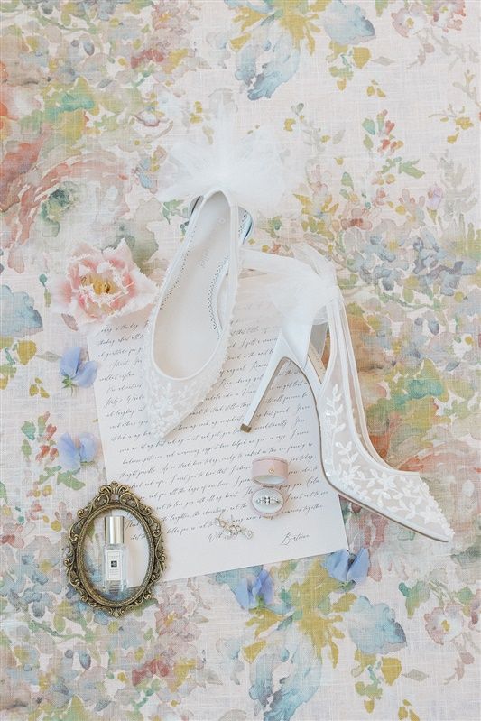 Elegant white lace wedding shoes on handwritten letter with antique cameo brooch against floral wallpaper background.