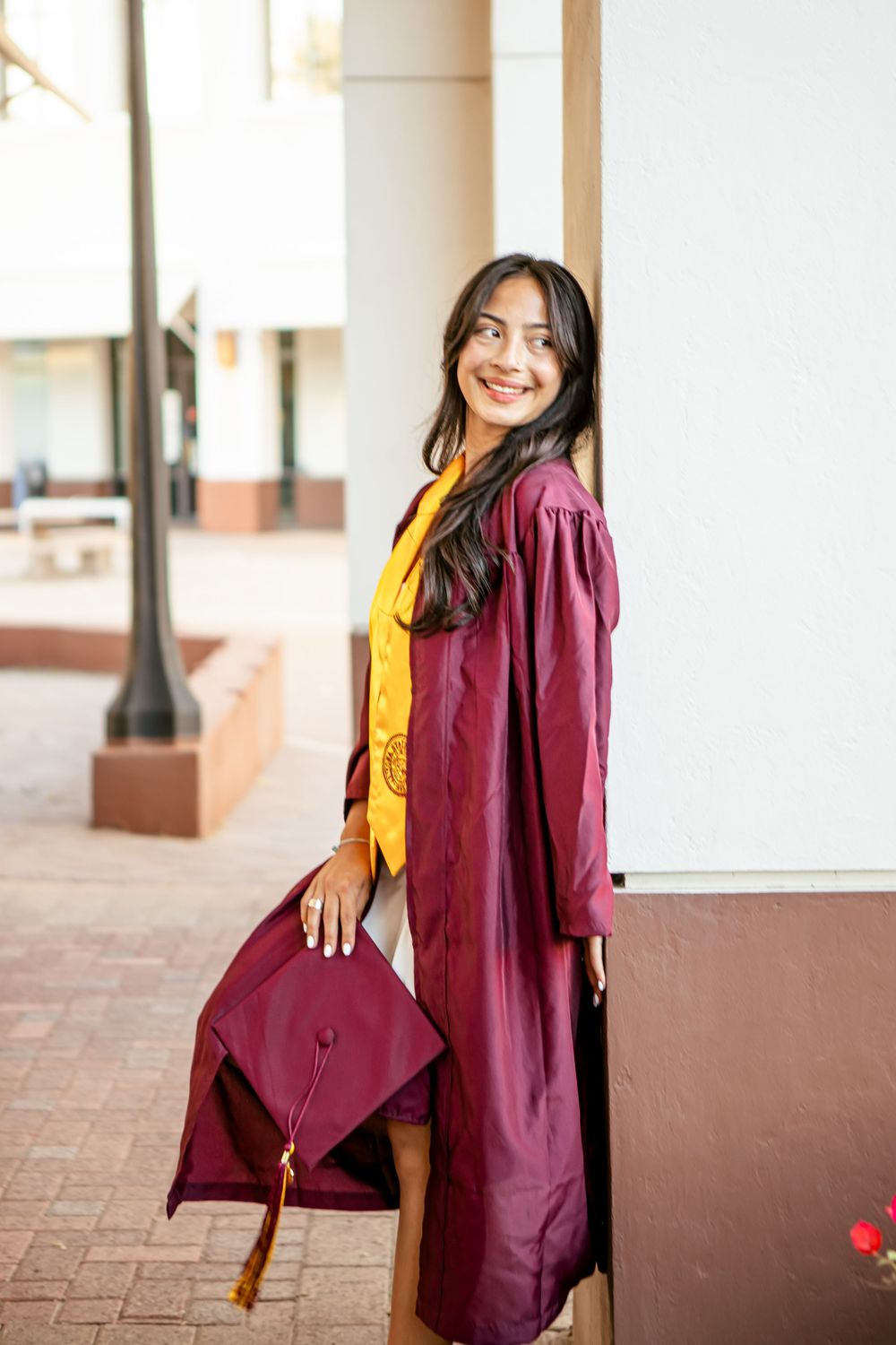Graduate in burgundy cap and gown poses against white building columns.