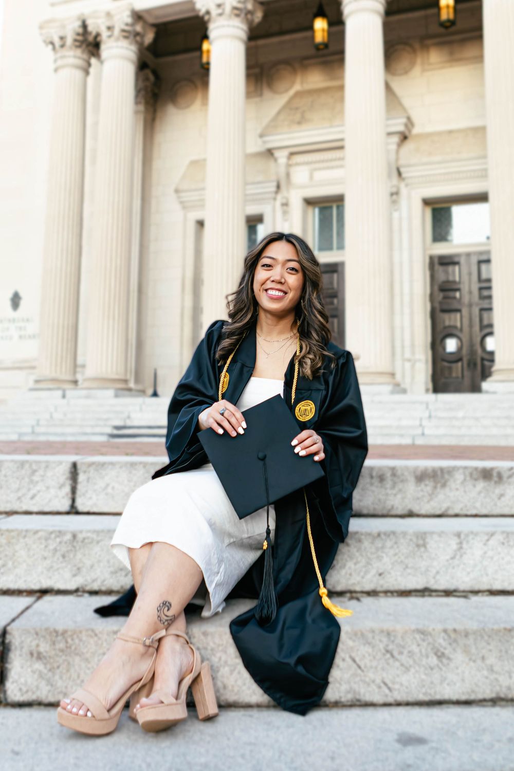 Smiling graduate in black cap and gown sits on university steps holding mortarboard, wearing white dress and heels.