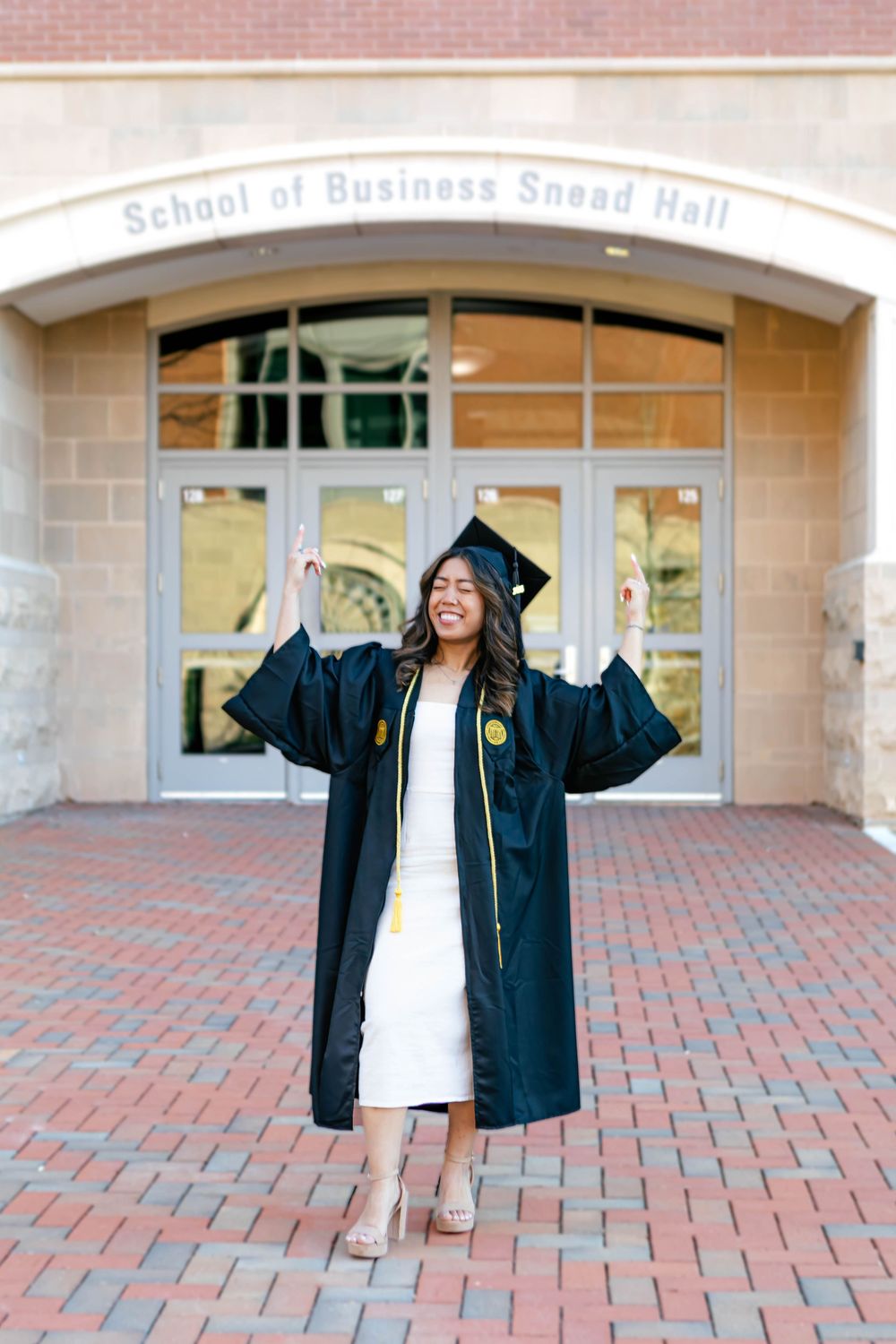 Smiling graduate in black cap and gown celebrates in front of School of Business Snead Hall on brick plaza.